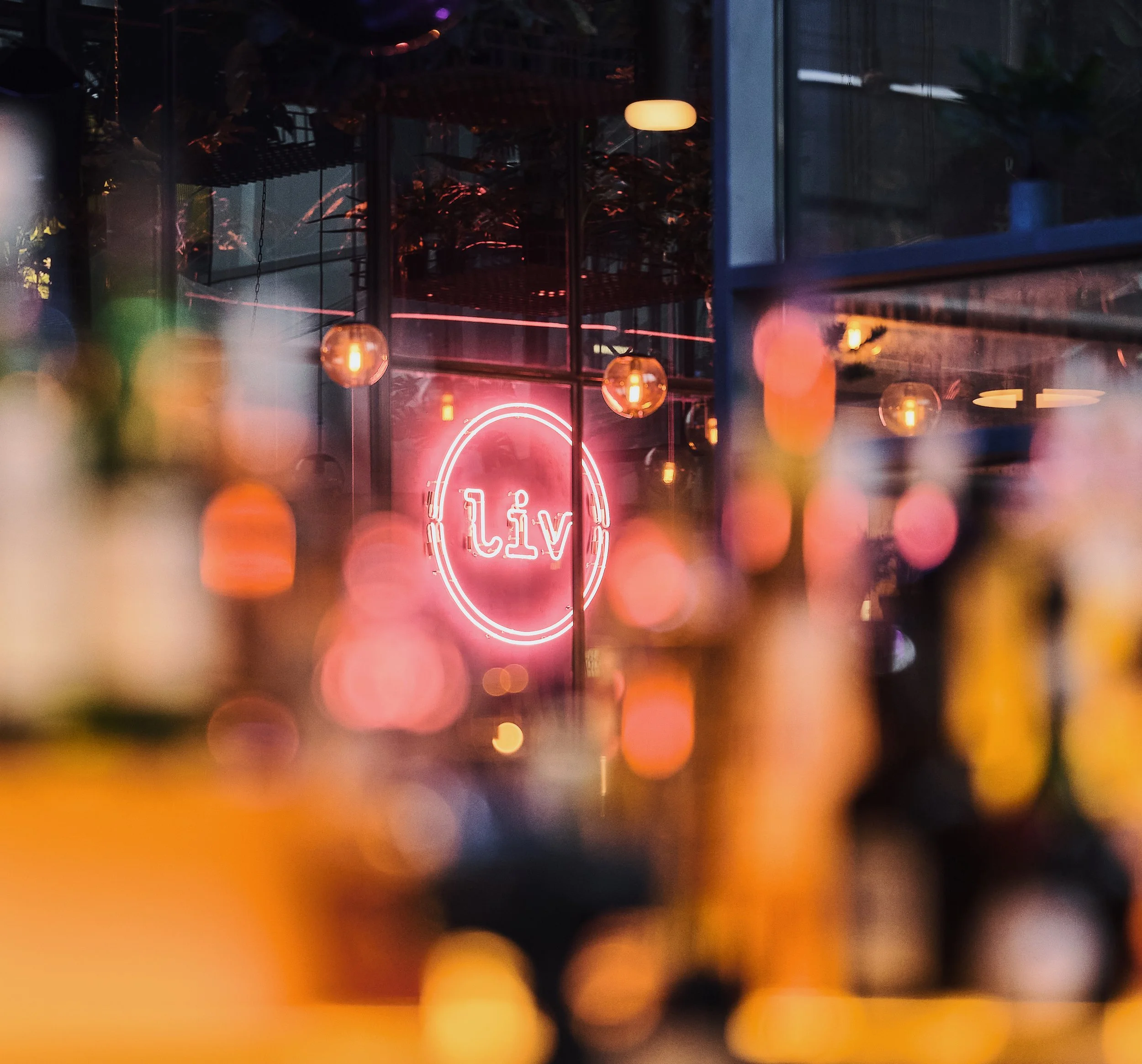 Liv Bar Bielefeld. Neon pink sign with the word 'Liv' inside a circle in a dimly lit interior, with warm hanging lights and reflections on windows, creating a vibrant and cozy atmosphere.