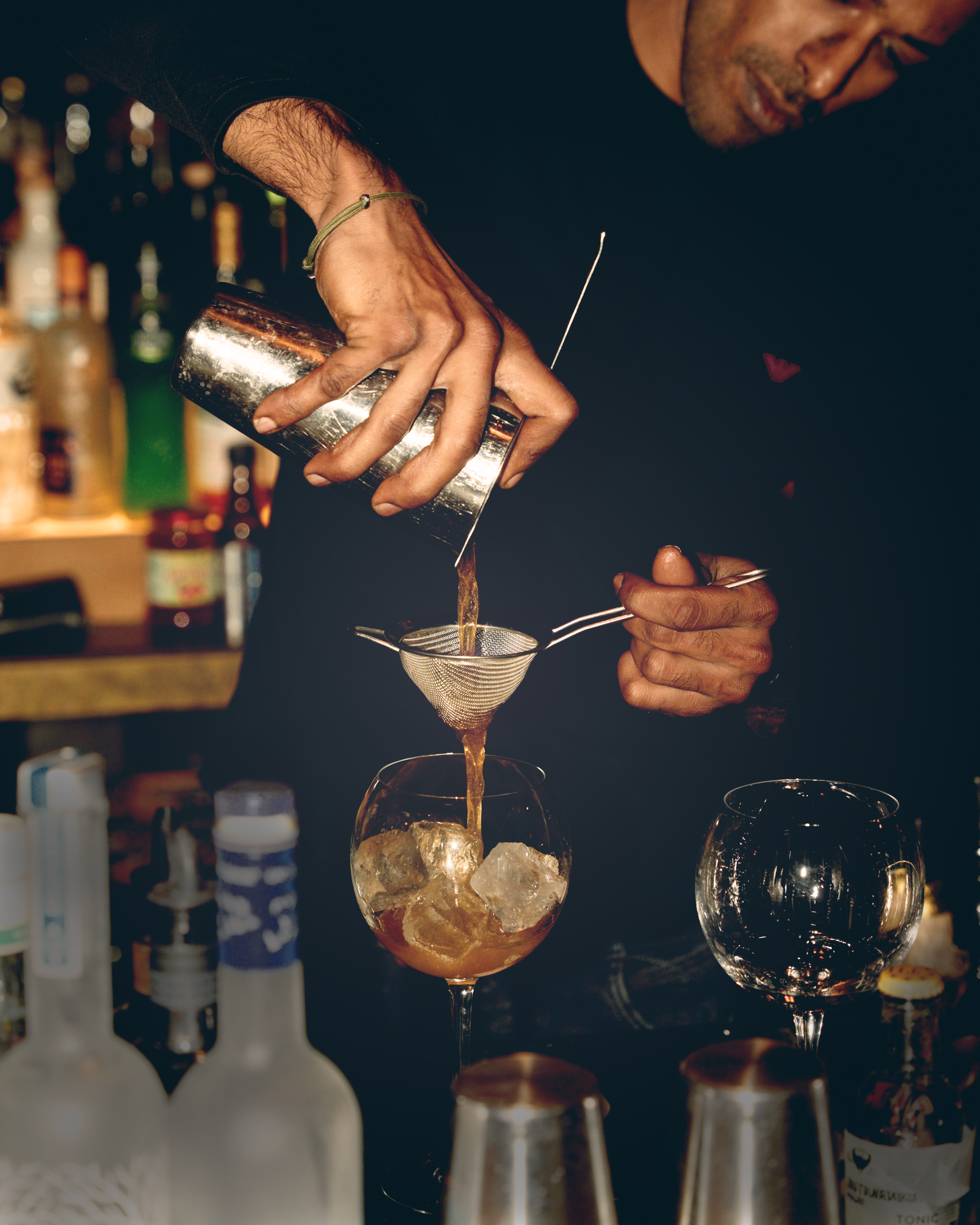 Liv Bar Bielefeld. Bartender pours liquid through a mesh strainer into a cocktail glass filled with ice at a bar.
