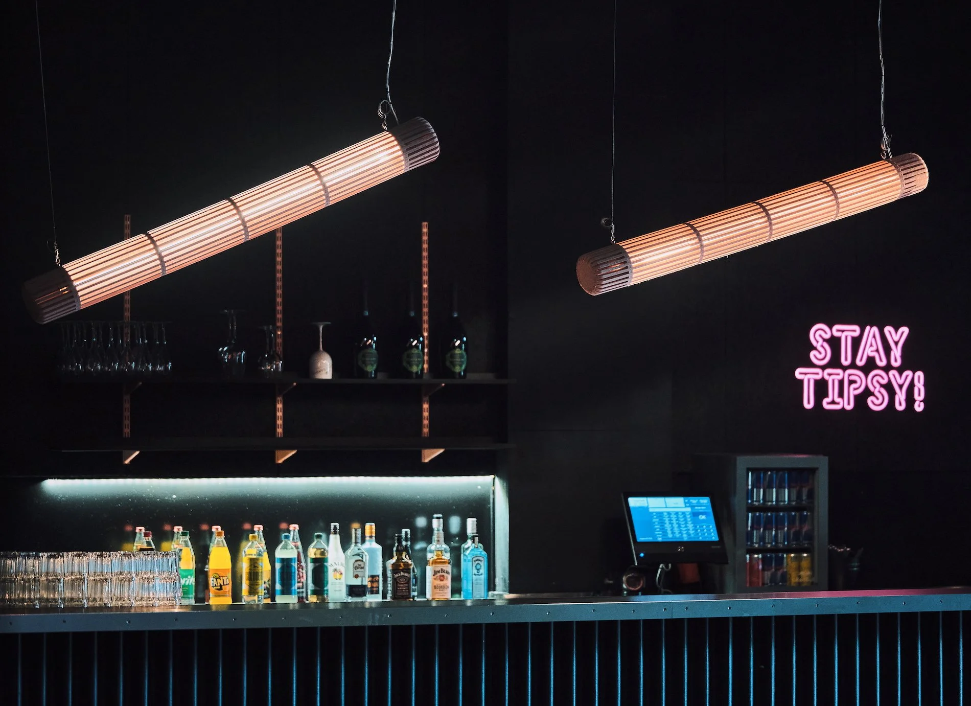 Stereo Bielefeld Club. A bar with bottles of liquor on the counter, glasses on the shelf, and two hanging wood light fixtures. A pink neon sign says 'STAY TIPSY' on the dark wall.