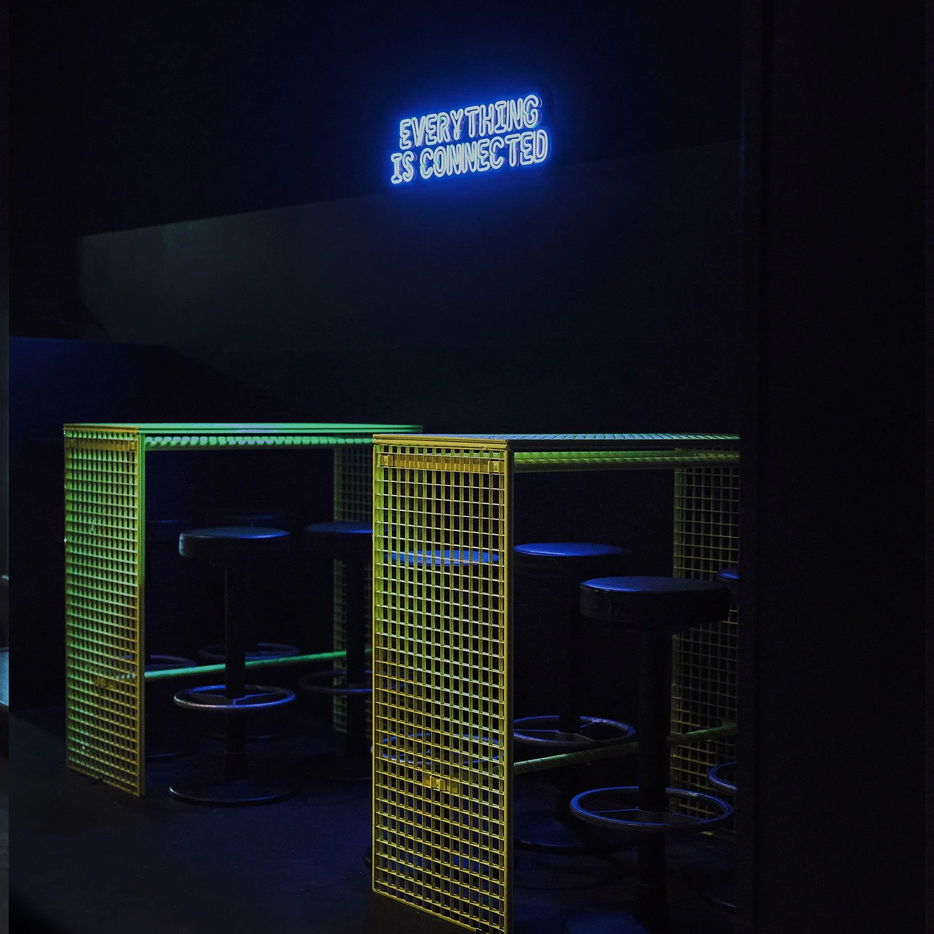 Stereo Bielefeld Club. Dark room with neon sign reading 'Everything is connected' and a yellow and green metal fence structure with black bar stools underneath.