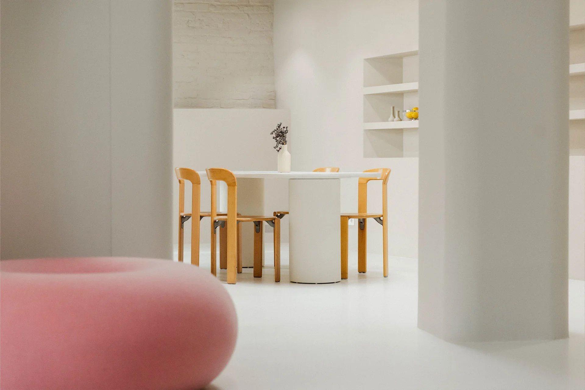 Studio fiveOh Köln. Haptik Agentur. Modern minimalist dining area with a white round table, four wooden chairs, a white vase with a small plant, and a white shelf with decorative items, in a bright room with white walls and a light-colored floor.