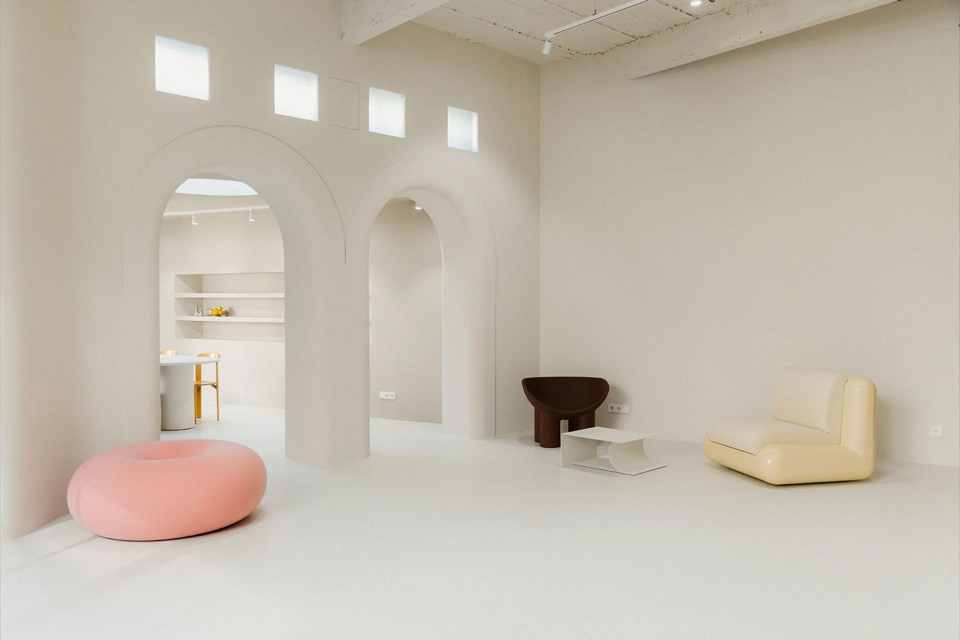 Studio fiveOh Köln. Haptik Agentur. Minimalist interior of a room with white walls and floor, featuring modern furniture including a pink oval ottoman, a brown curved chair, a beige curved sofa, and a white side table. 