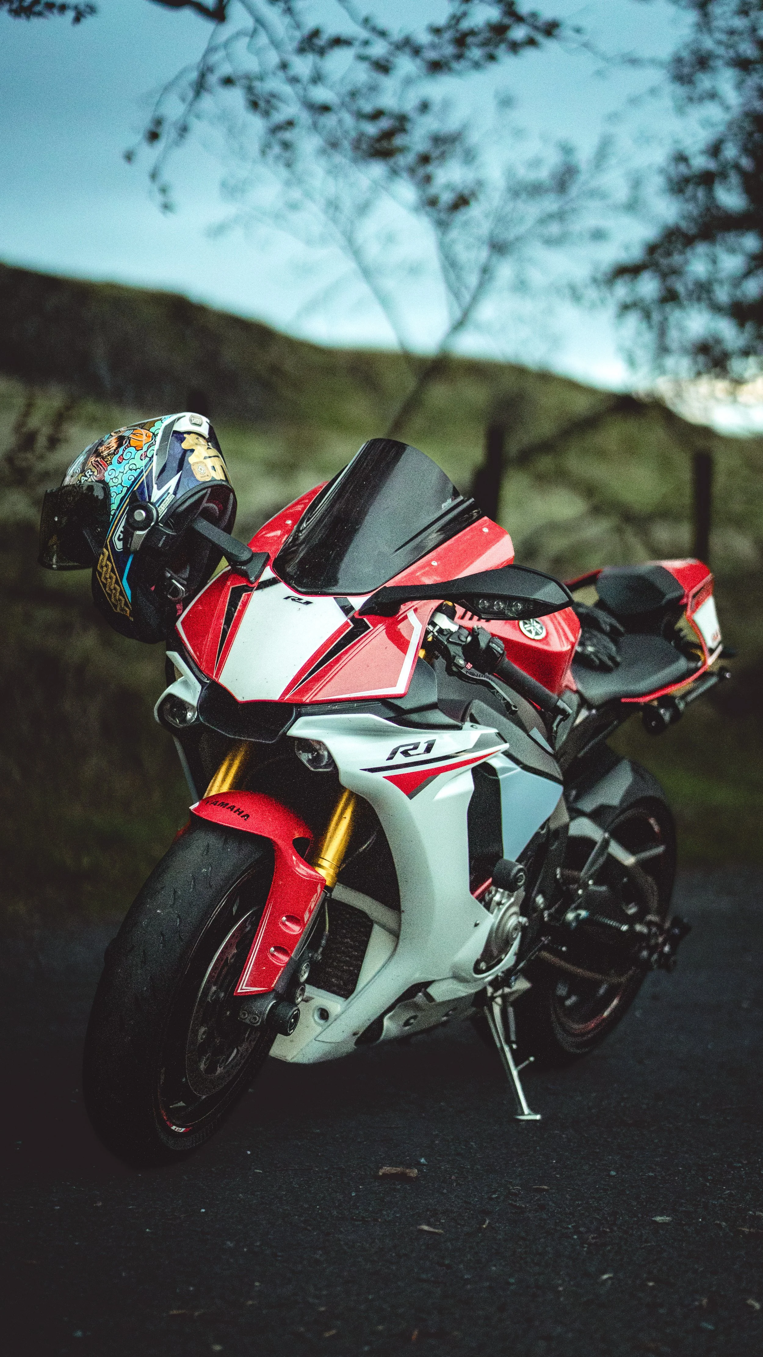 A Yamaha YZF-R1 motorcycle parked on a road with a helmet hanging on its handlebar. The background features blurry trees and rolling hills.