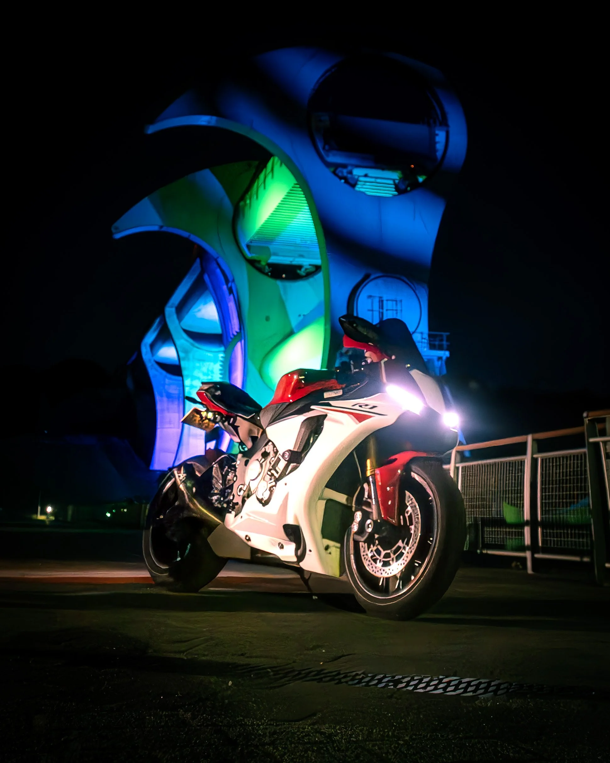 A white and red sport motorcycle parked at night in front of a colorful, illuminated building with organic, flowing shapes.
