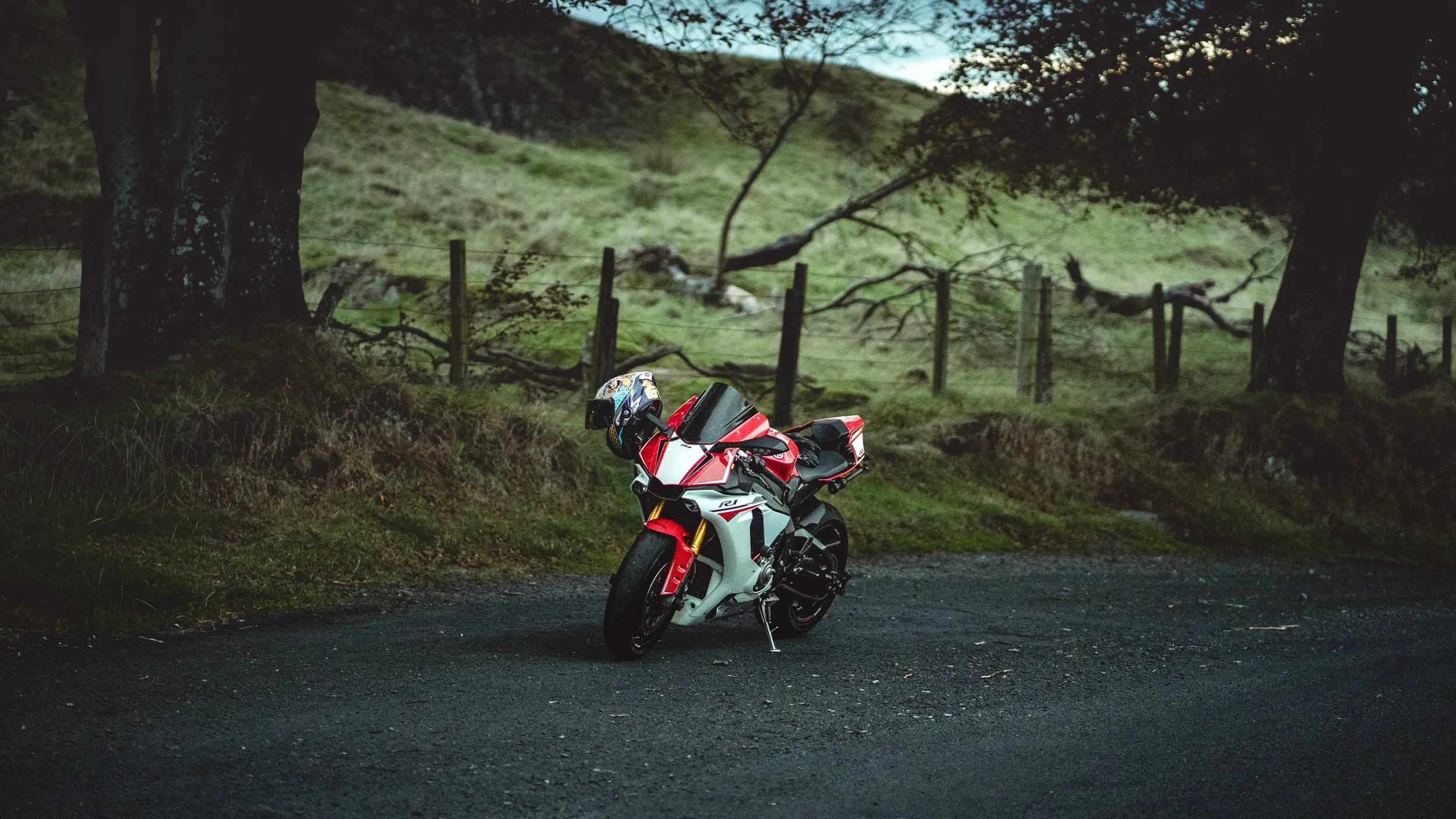 A red and white sport motorcycle parked on a dark, gravel road in a rural area with grassy hills, trees, and a fence in the background.