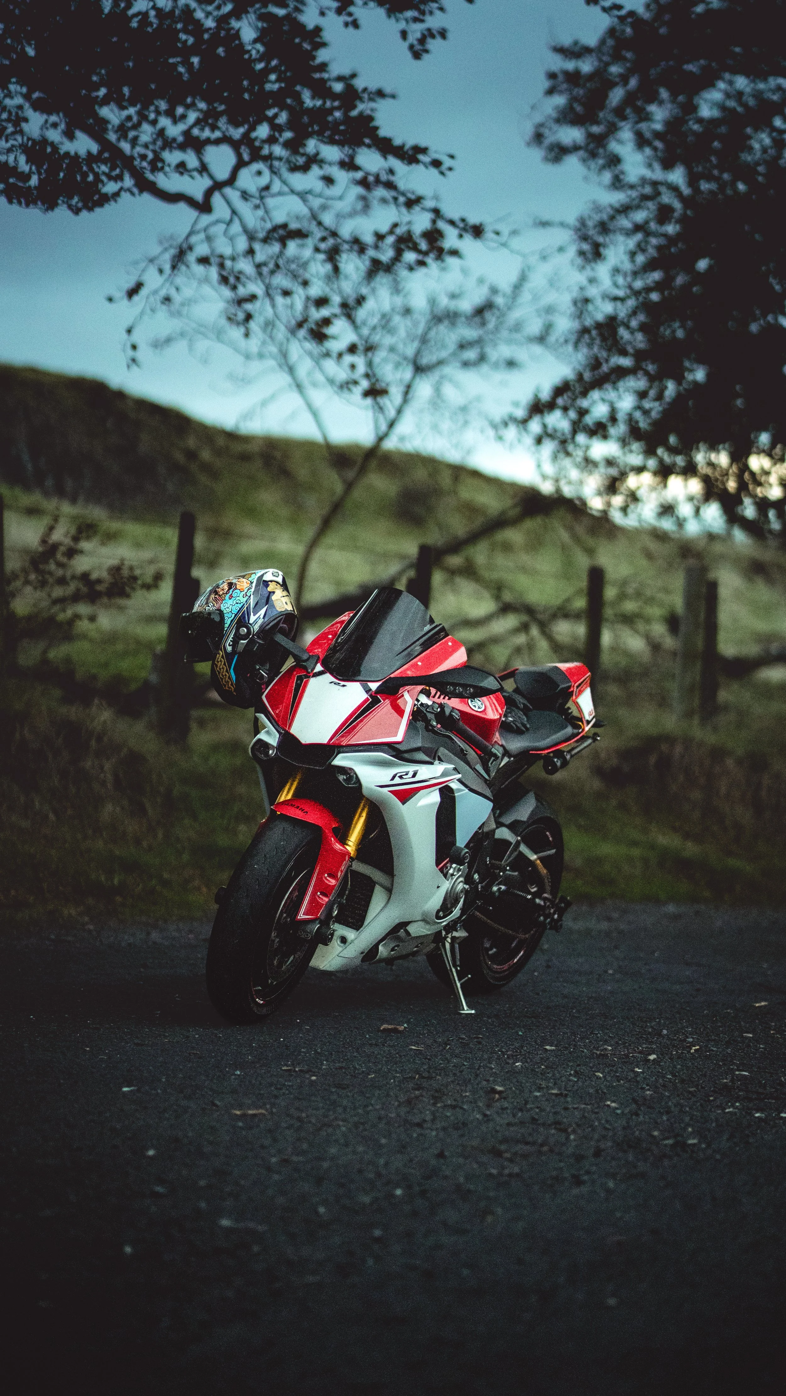 A red, white, and black sport motorcycle parked on a dark road in a rural area with a helmet hanging on its handlebar, trees, and gently rolling hills in the background during dusk.