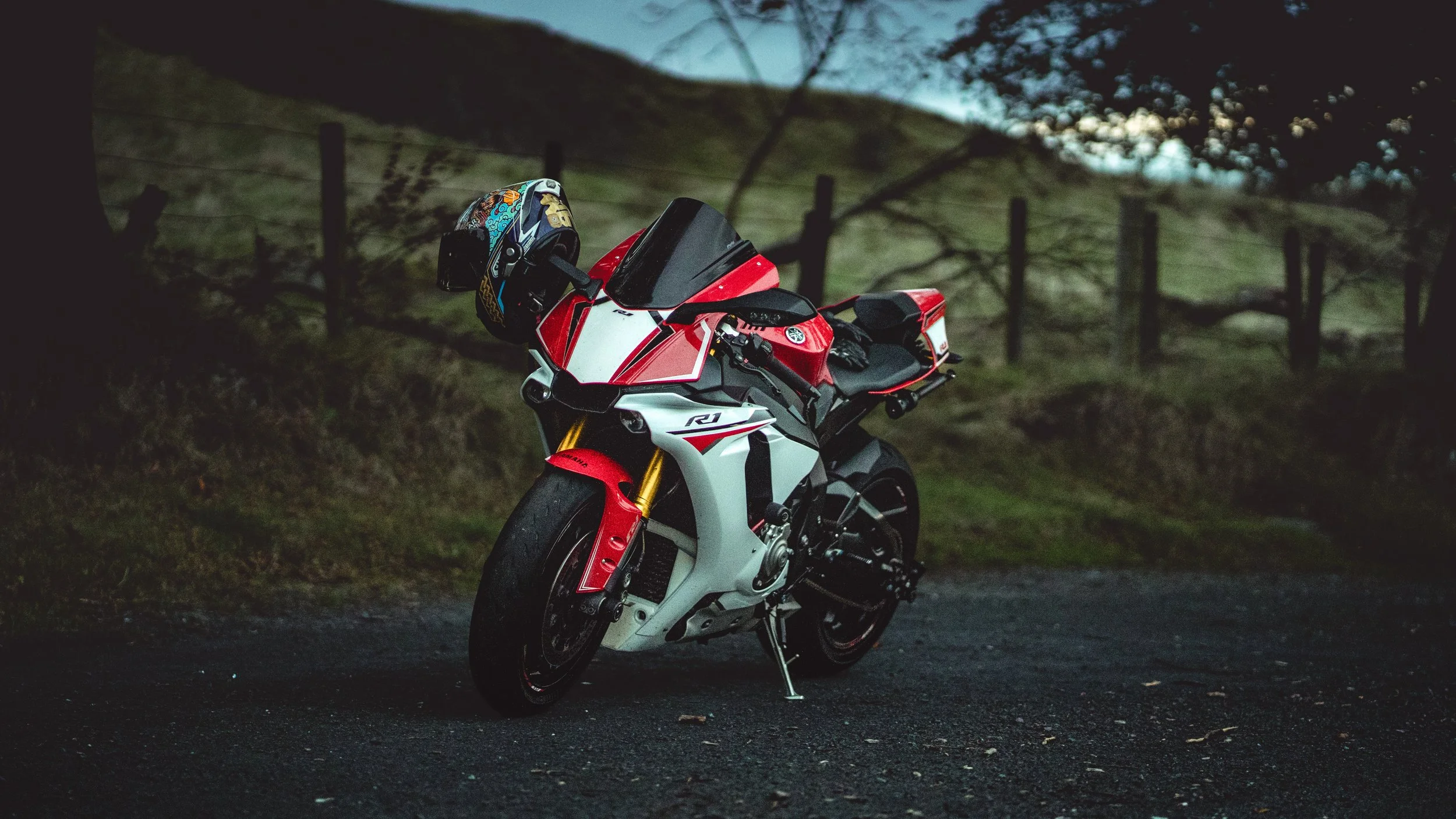 Red and white Yamaha R1 sport motorcycle parked on a dark road at dusk, with a colorful helmet resting on the handlebars and a grassy hillside with trees and a fence in the background.