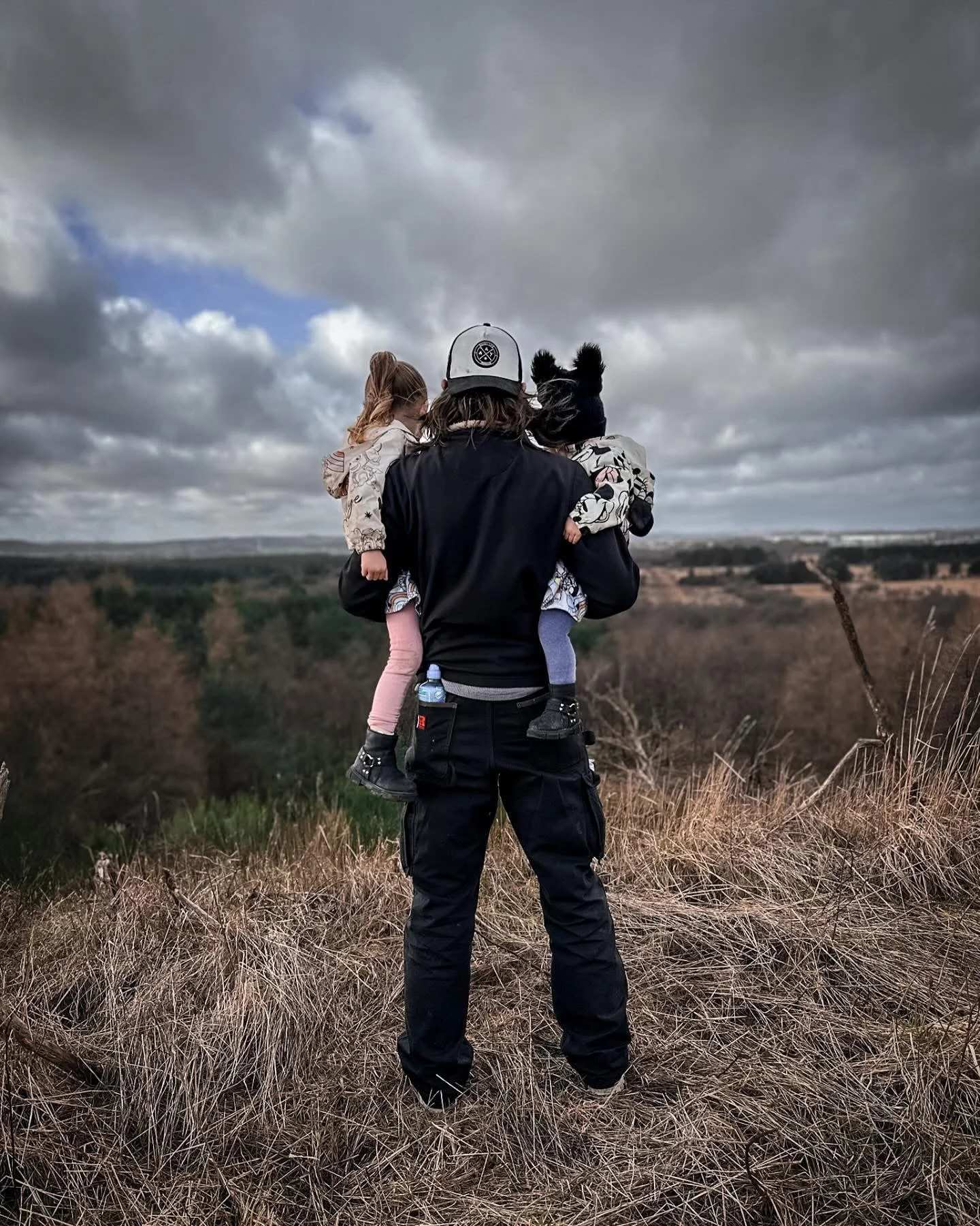 A person wearing a black jacket and cap holding two children, one on each side, standing on a grassy hill with a cloudy sky and distant landscape in the background.