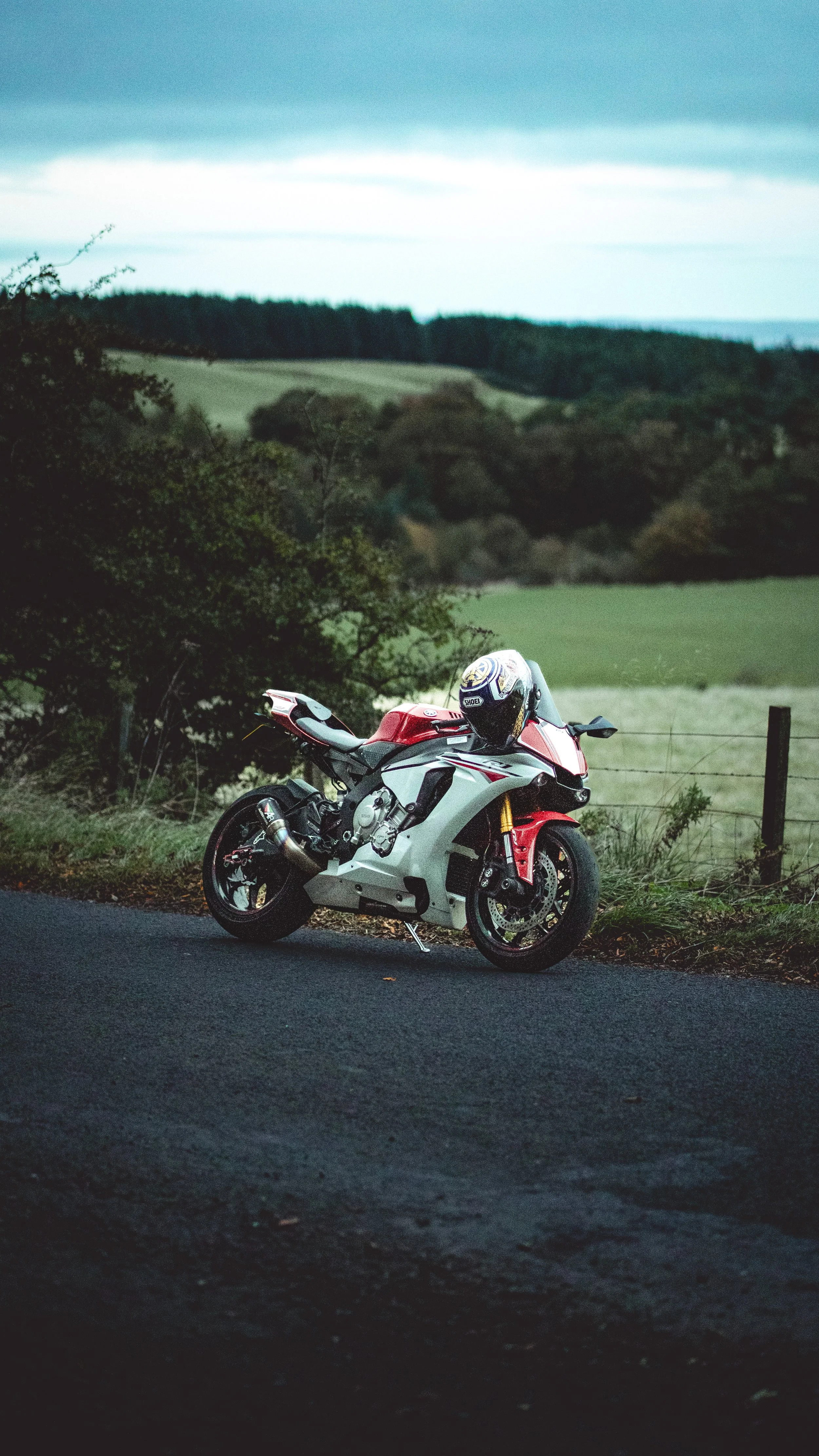 A red and white sports motorcycle with a helmet resting on the handlebars, parked on the side of a rural road with green fields and rolling hills in the background.