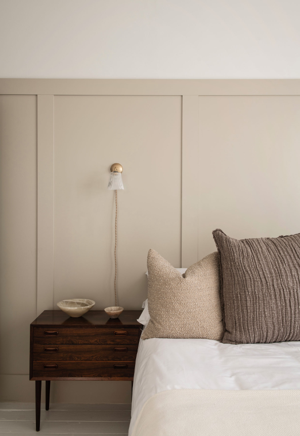 Close-up of a bedroom with a beige headboard, two pillows, and a wooden nightstand holding ceramic bowls, with a wall-mounted lamp above.
