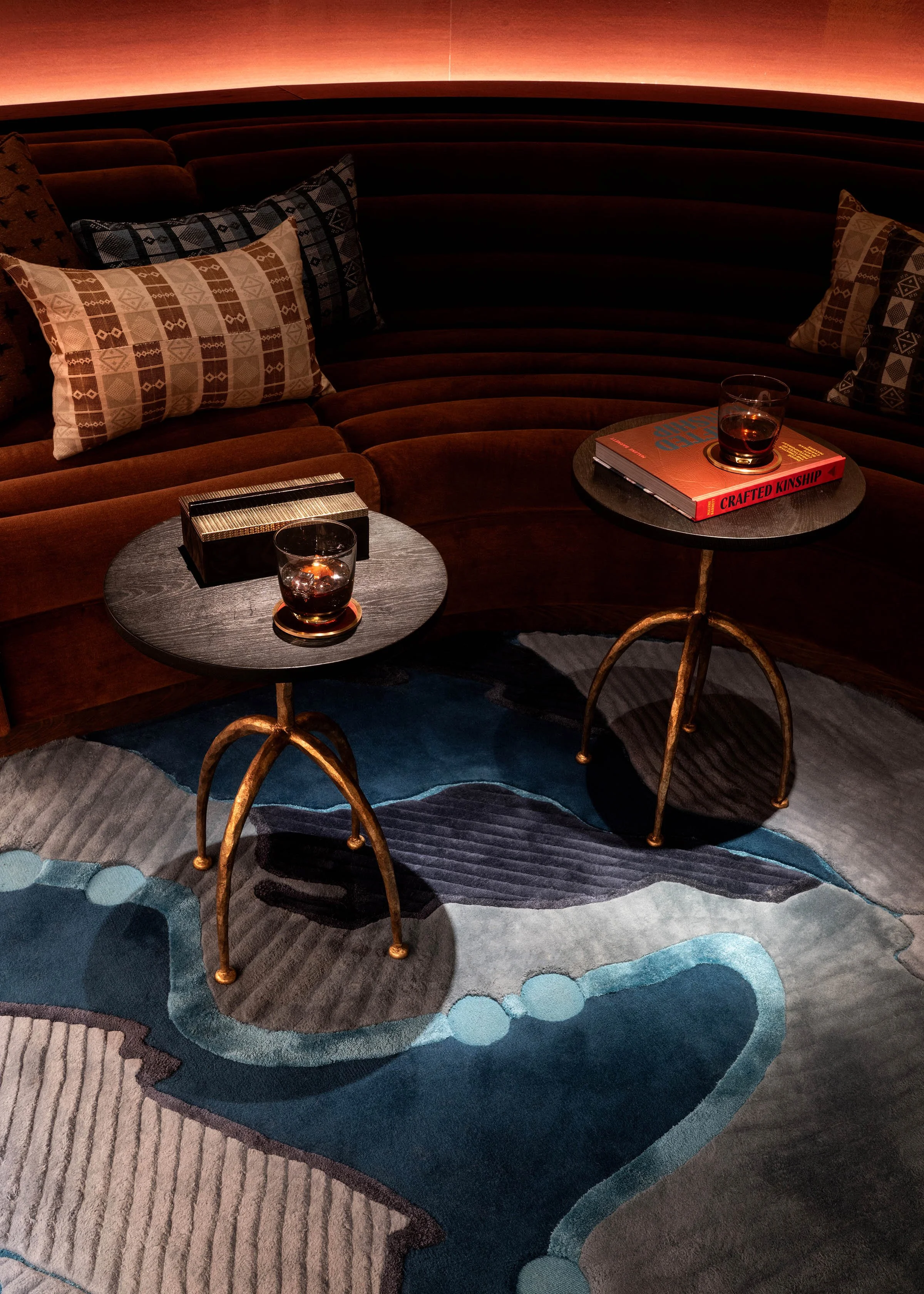 A cozy seating area with a curved, dark brown velvet sofa adorned with patterned pillows, two round black tables with gold legs holding glasses of dark beverages and books; an abstract multicolored rug, and warm ambient lighting in the background.