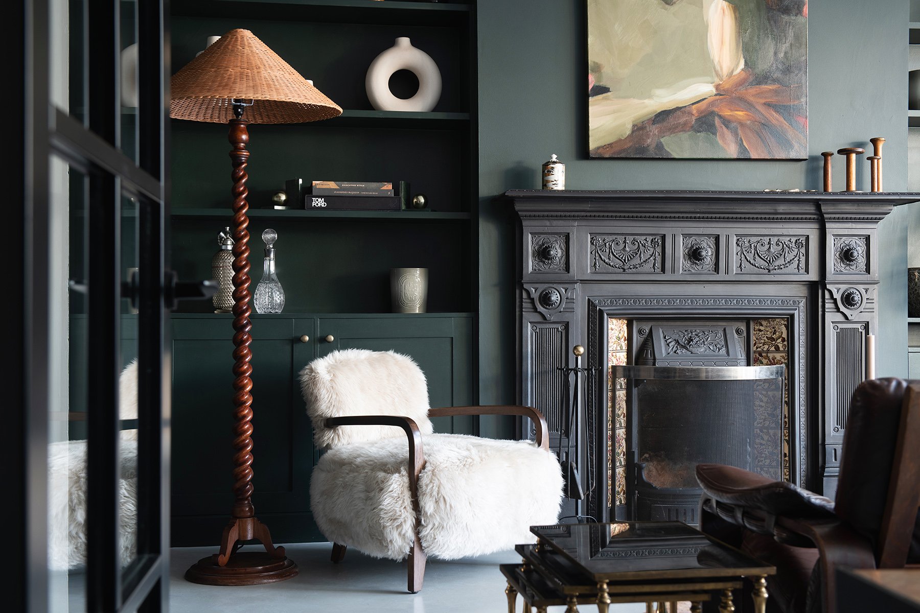 Interior of a living room with a dark green wall, a black fireplace with ornate detailing, a sheepskin armchair, a wooden floor lamp with a woven shade, paintings, vases, and books on the wall shelves.