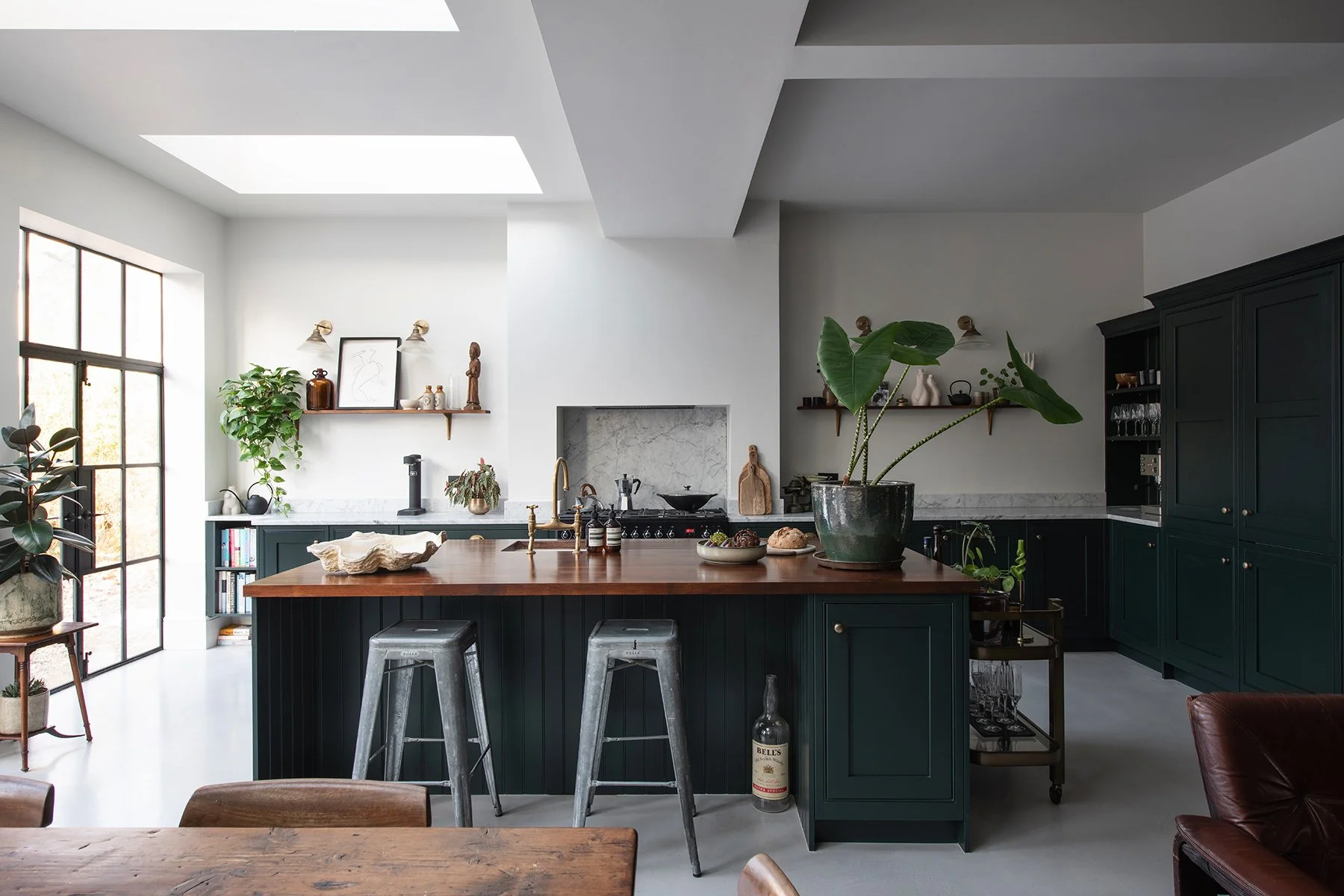 Modern kitchen with dark green cabinets, wooden island with bar stools, large window, plants, and decorative items