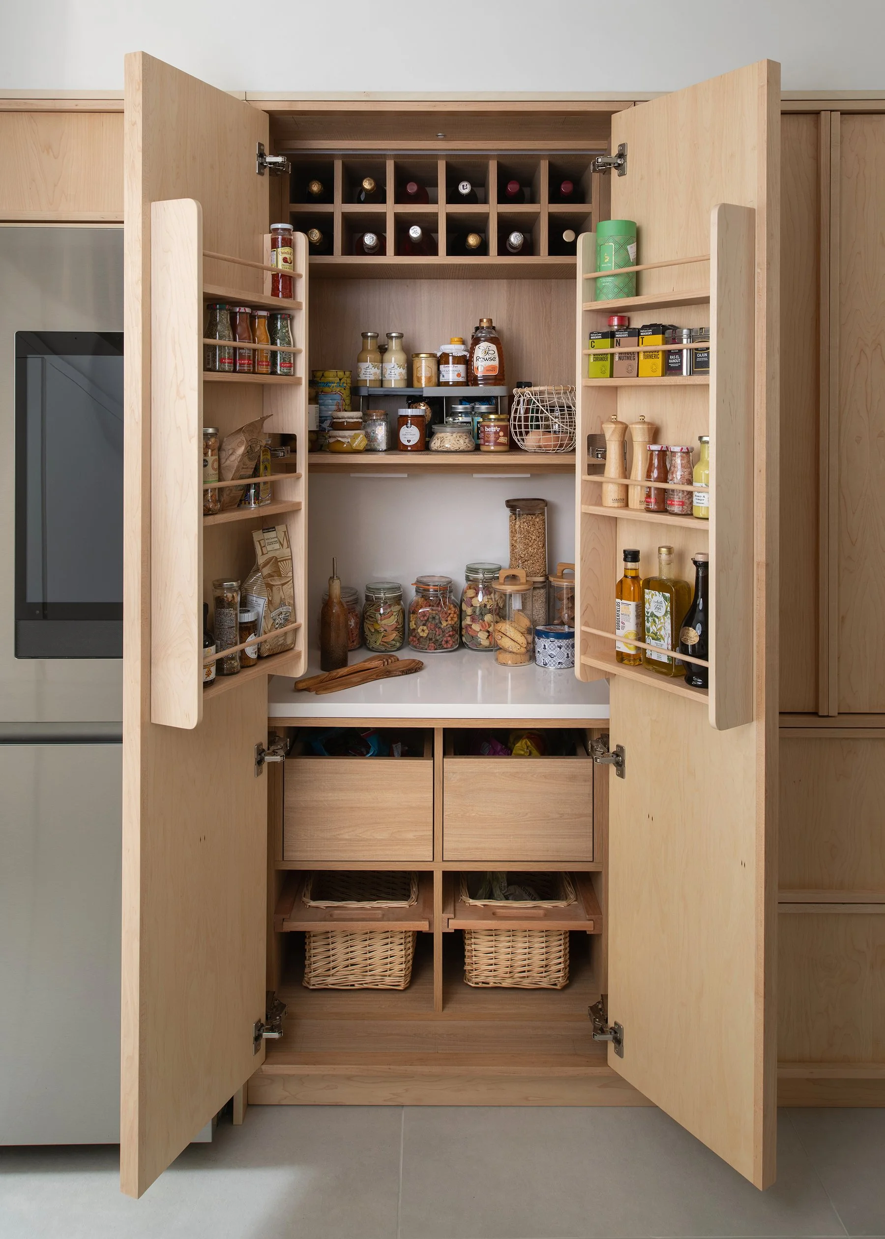Open kitchen cabinet with various shelves filled with jars, bottles, and containers of food and spices, with baskets at the bottom.