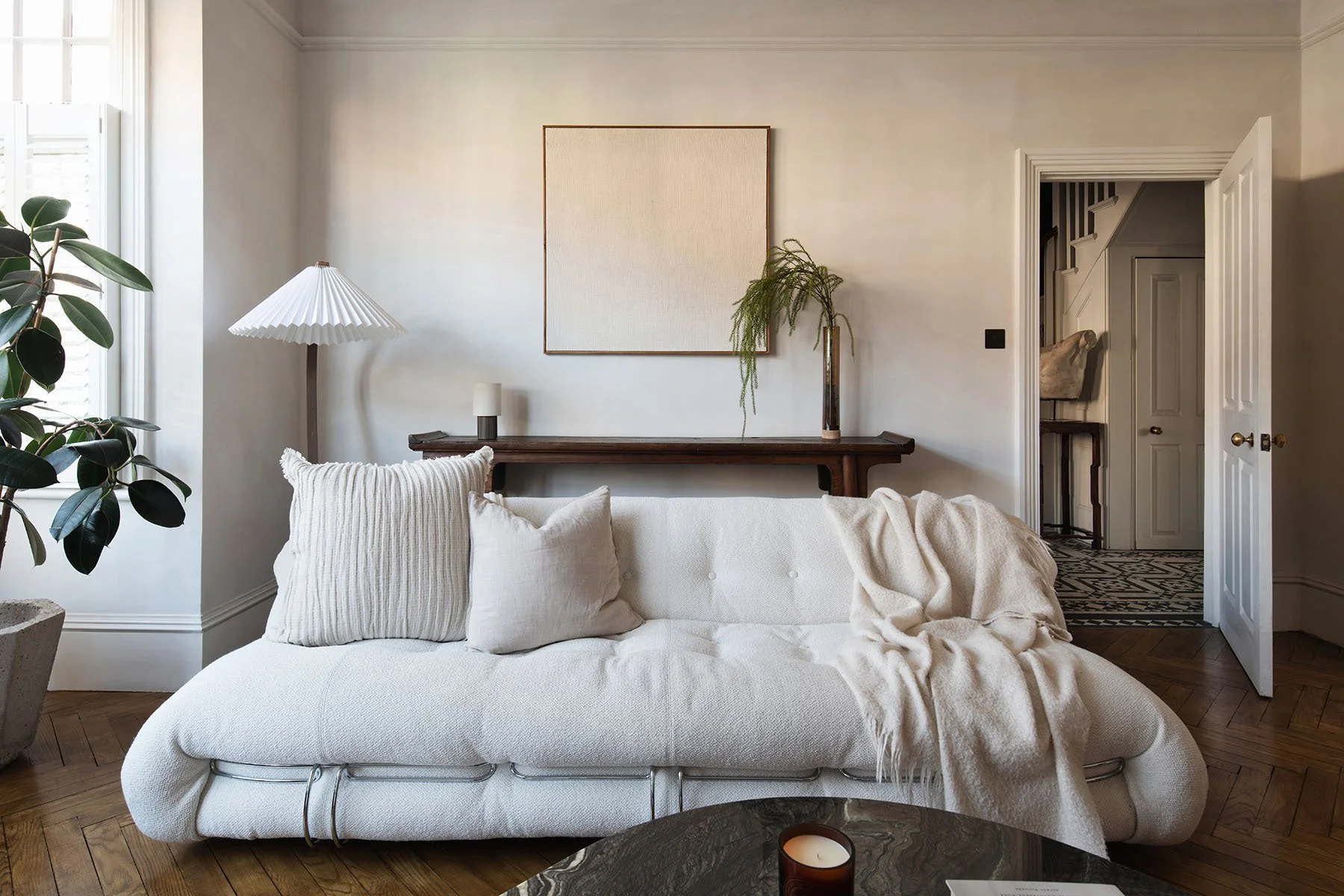 Living room with a white sofa, pillows, a blanket, a wooden floor, a potted plant, a large window, and a wall with art and decorative greenery.