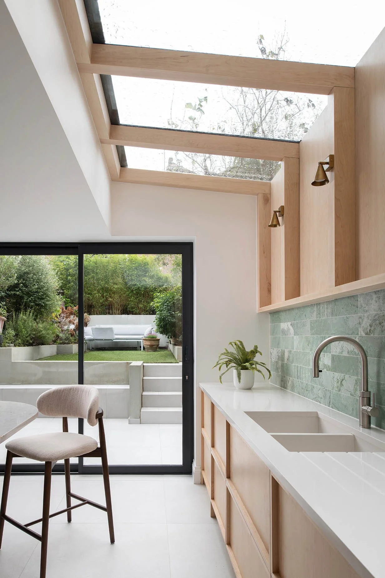 Modern kitchen with skylights, white countertops, green tiled backsplash, wooden cabinets, a potted plant, and sliding glass door leading to a garden patio with greenery and outdoor seating.