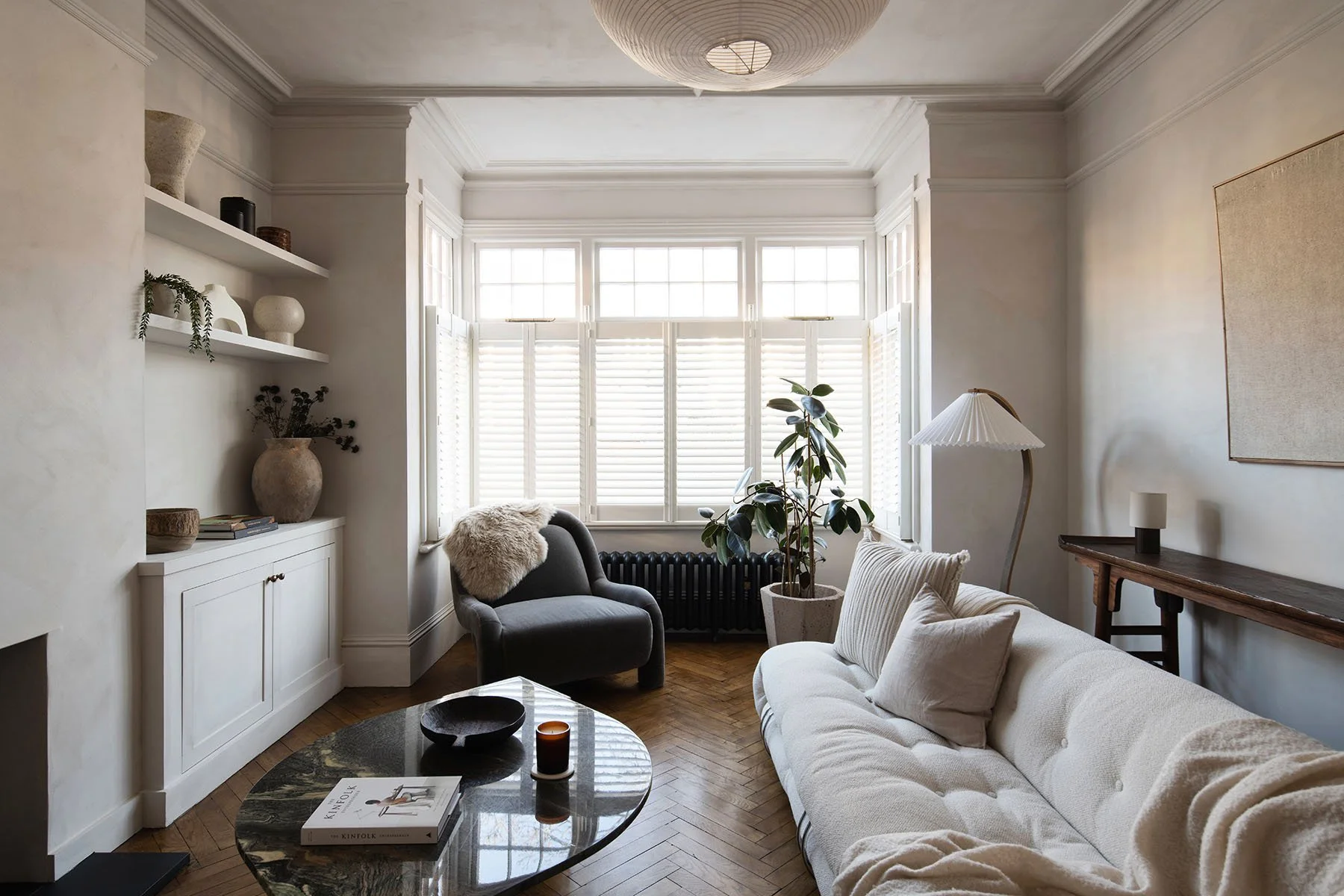 Living room with large window, white walls, and modern decor. Contains a white sofa, a dark armchair with a plush pillow, a floor lamp, a wooden console table with a small sculpture, a potted plant, and a coffee table with books and candles.
