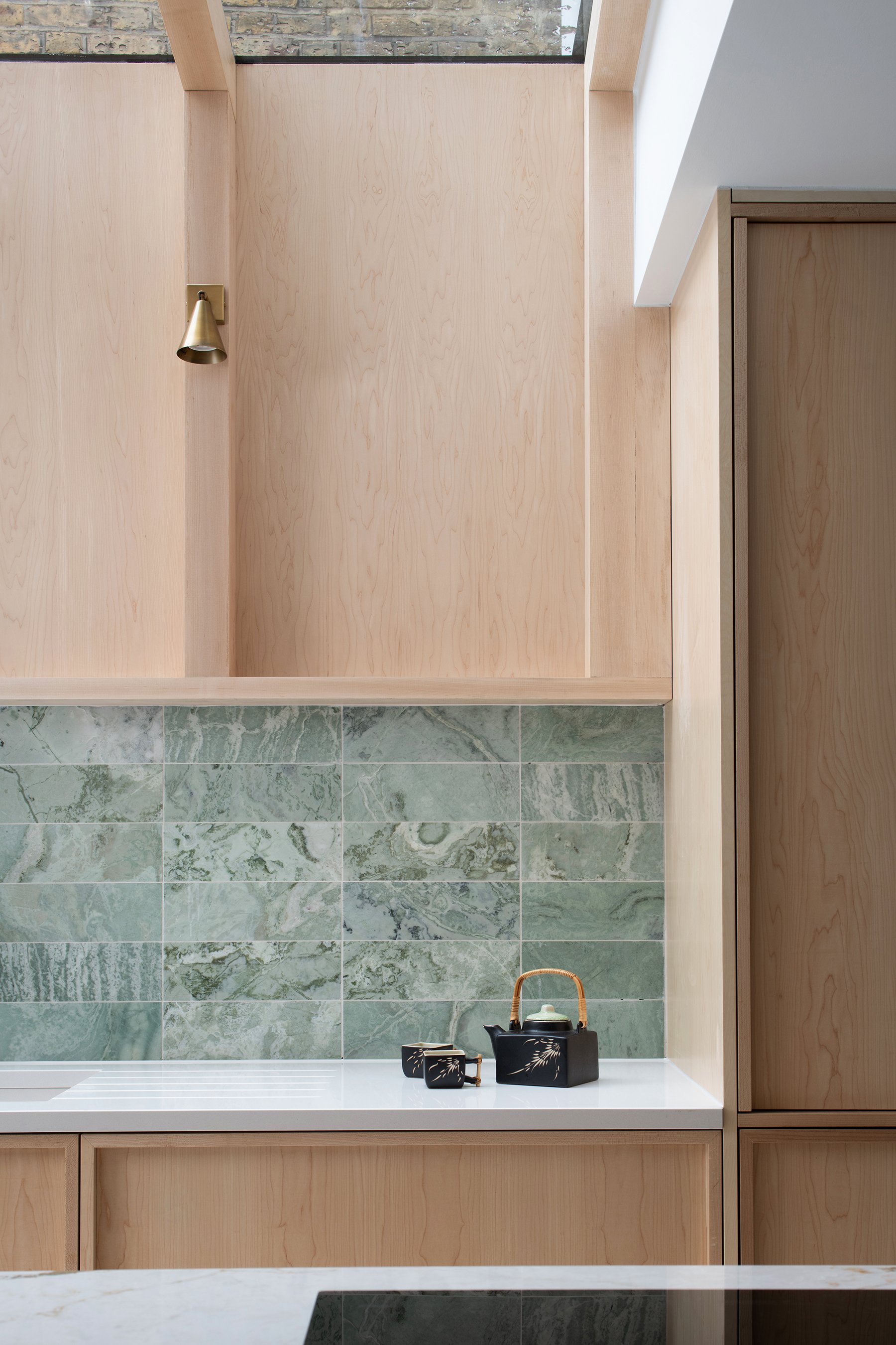 A minimalist kitchen with green marble backsplash, light wood cabinetry, and a white countertop featuring a black tea set with bamboo handle.
