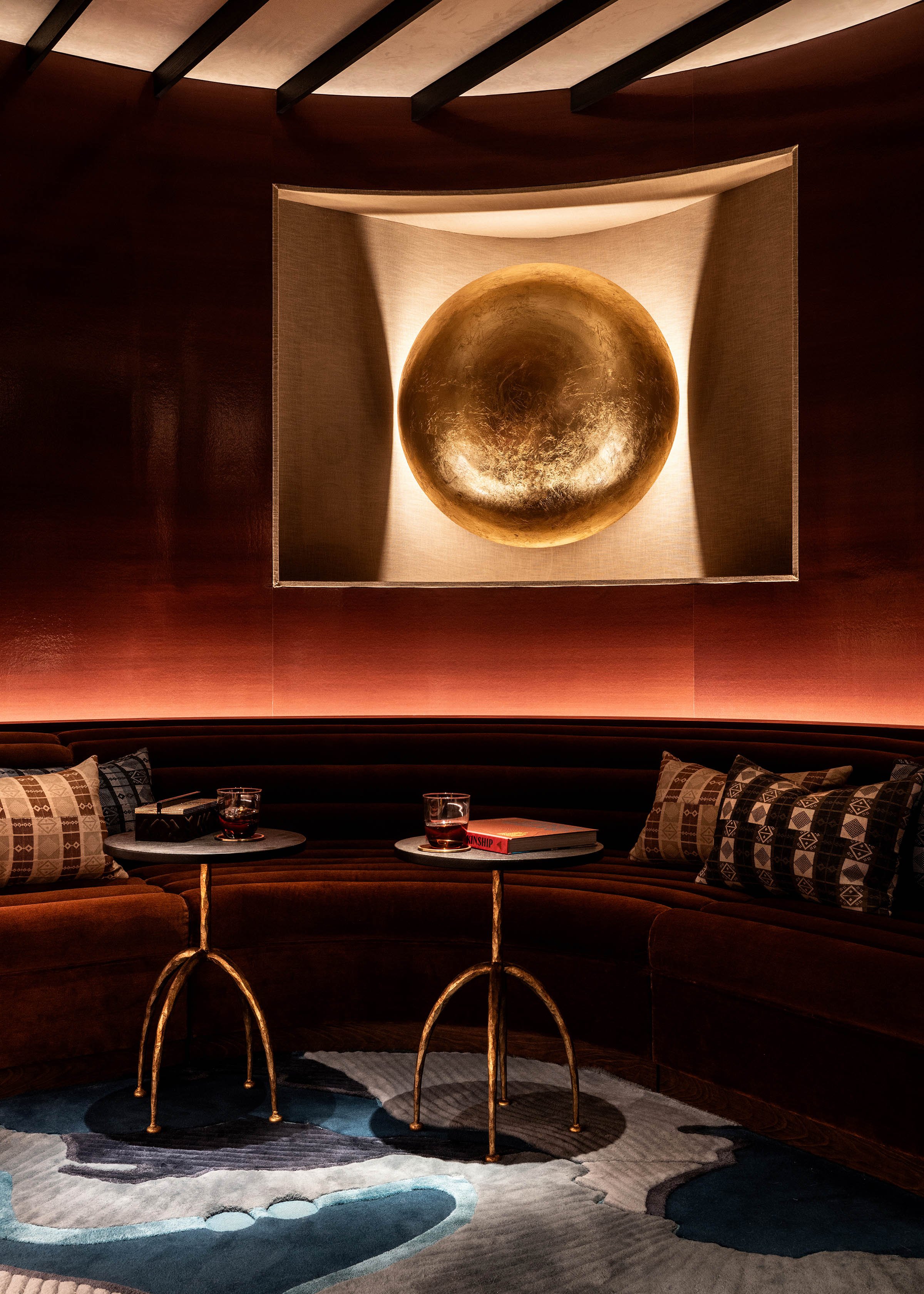 Luxurious seating area with curved dark brown velvet couch, decorative pillows, two small round tables with glasses of dark beverage, a book, and a remote, in front of a wall with a large, illuminated, gold spherical art piece.