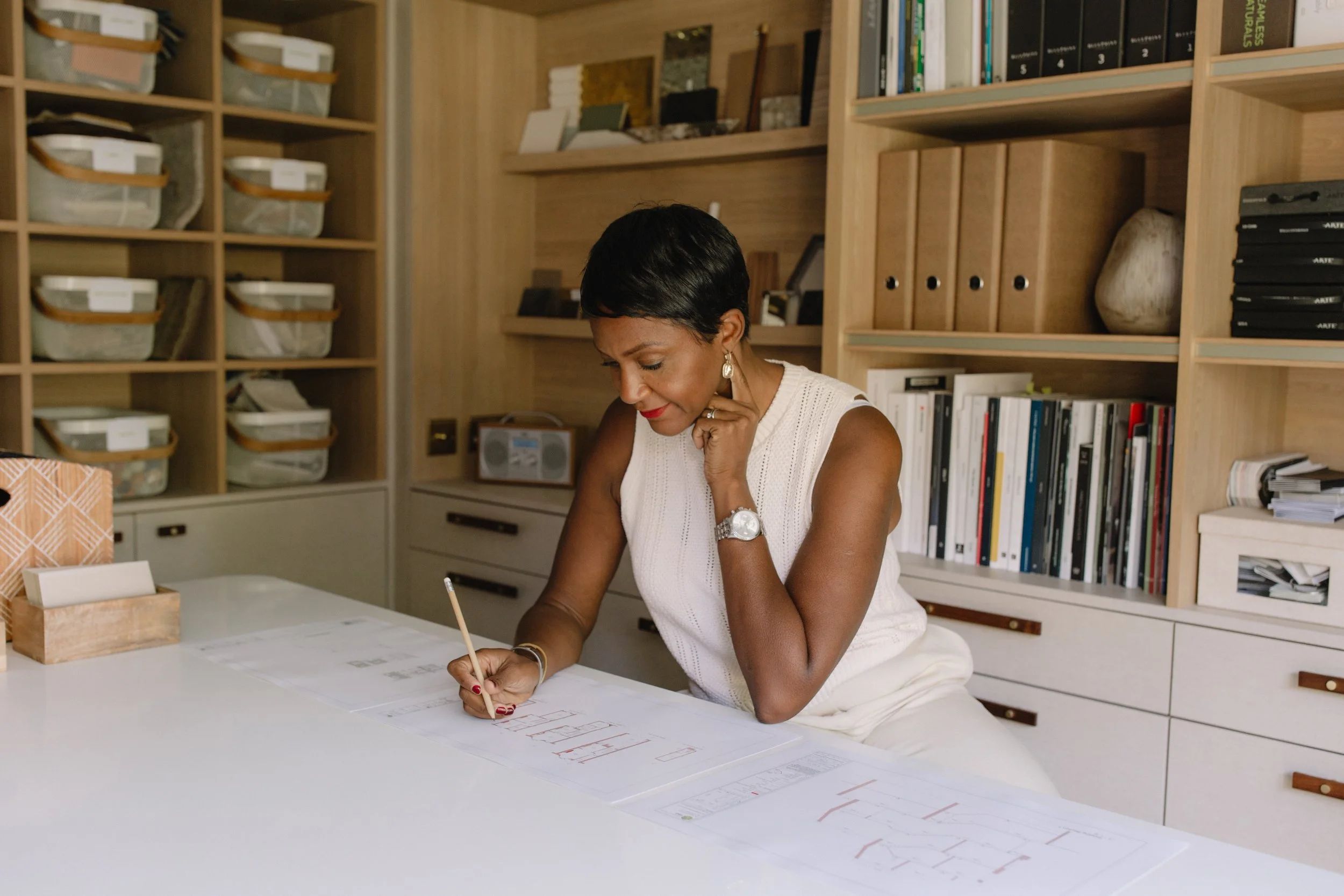 A woman sitting at a white table, working on architectural drawings with a pencil, in a room with wooden shelves filled with books, files, and decorative items.