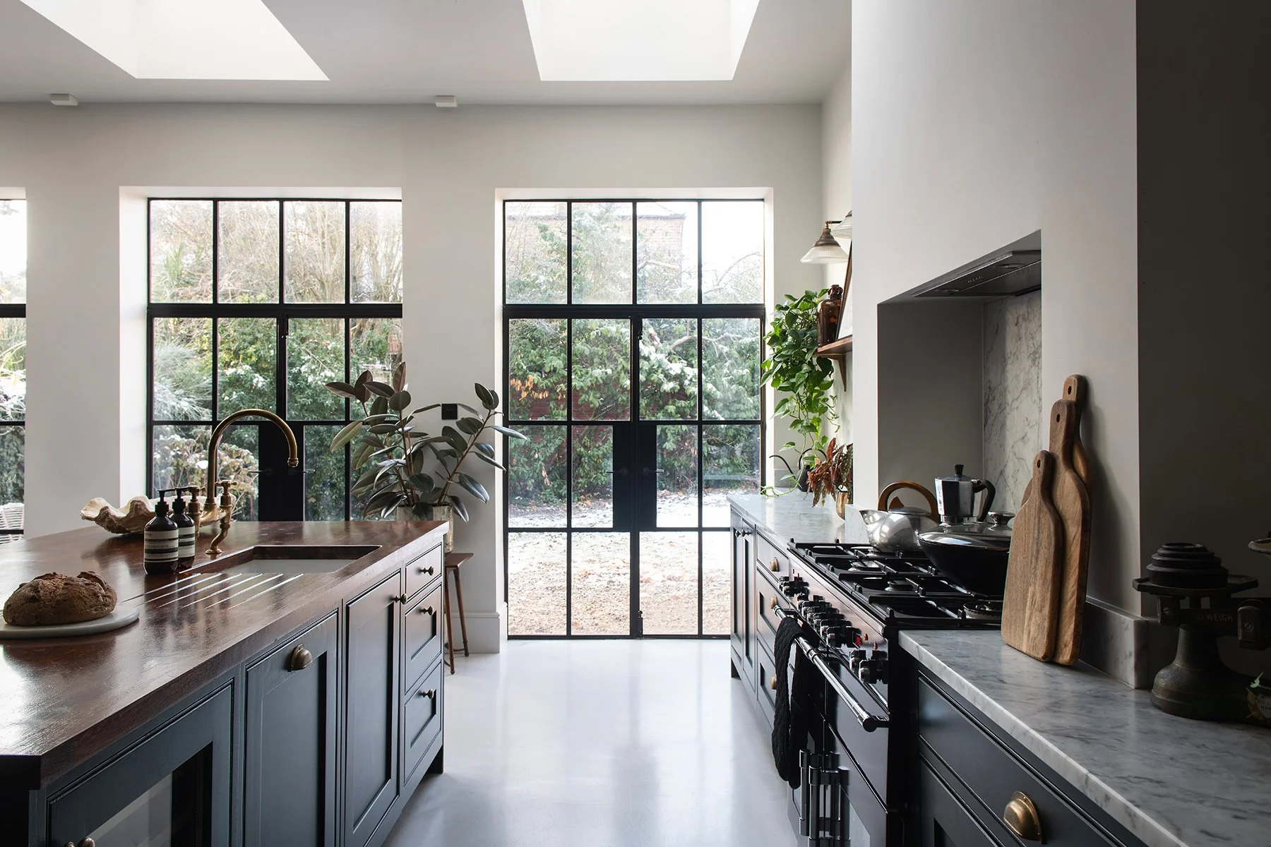 Modern kitchen with large windows overlooking a garden, featuring black cabinetry, a marble countertop, plants, and a wooden island with a sink.