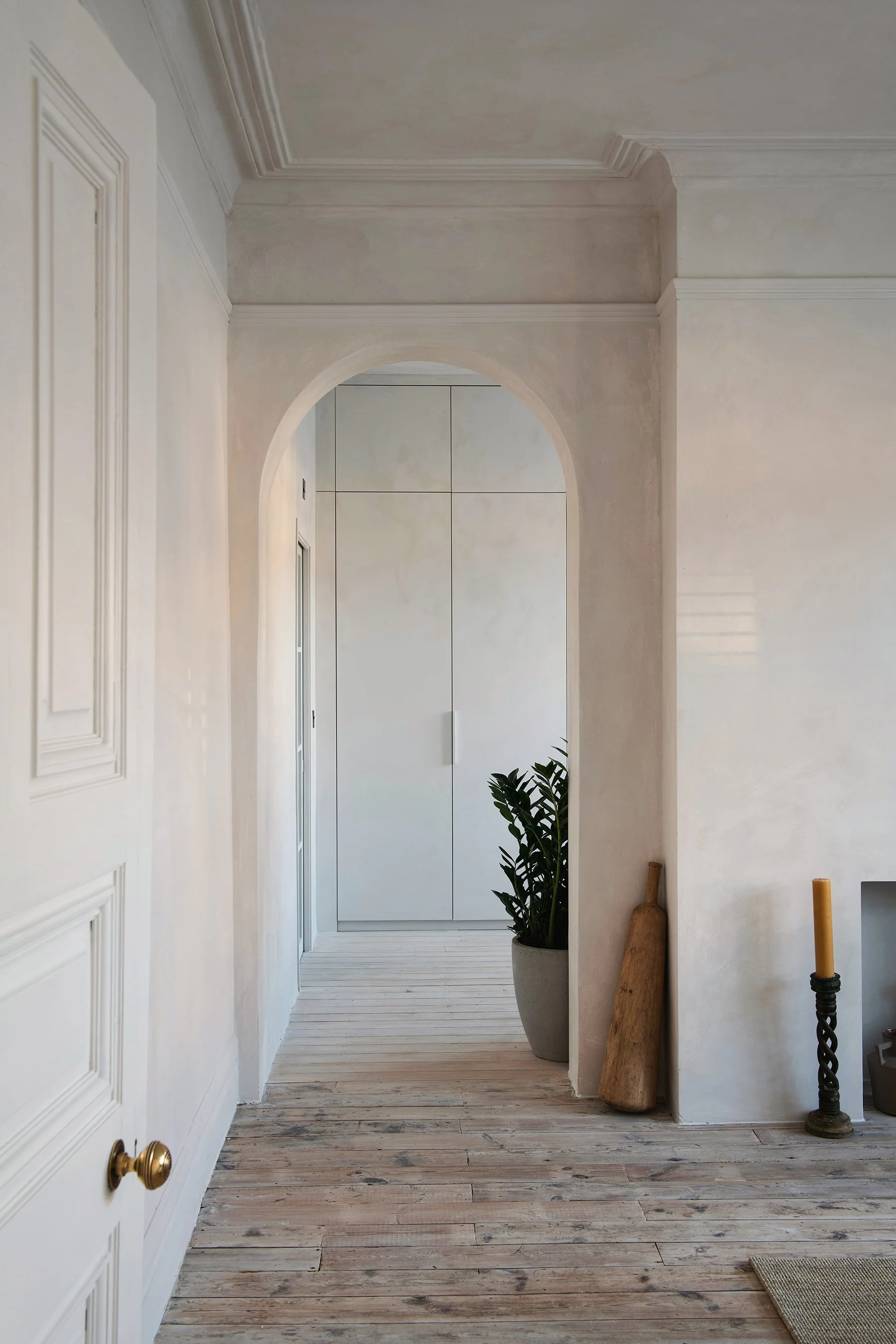 Entryway with white walls, wooden flooring, a potted plant, and tall candle holder.