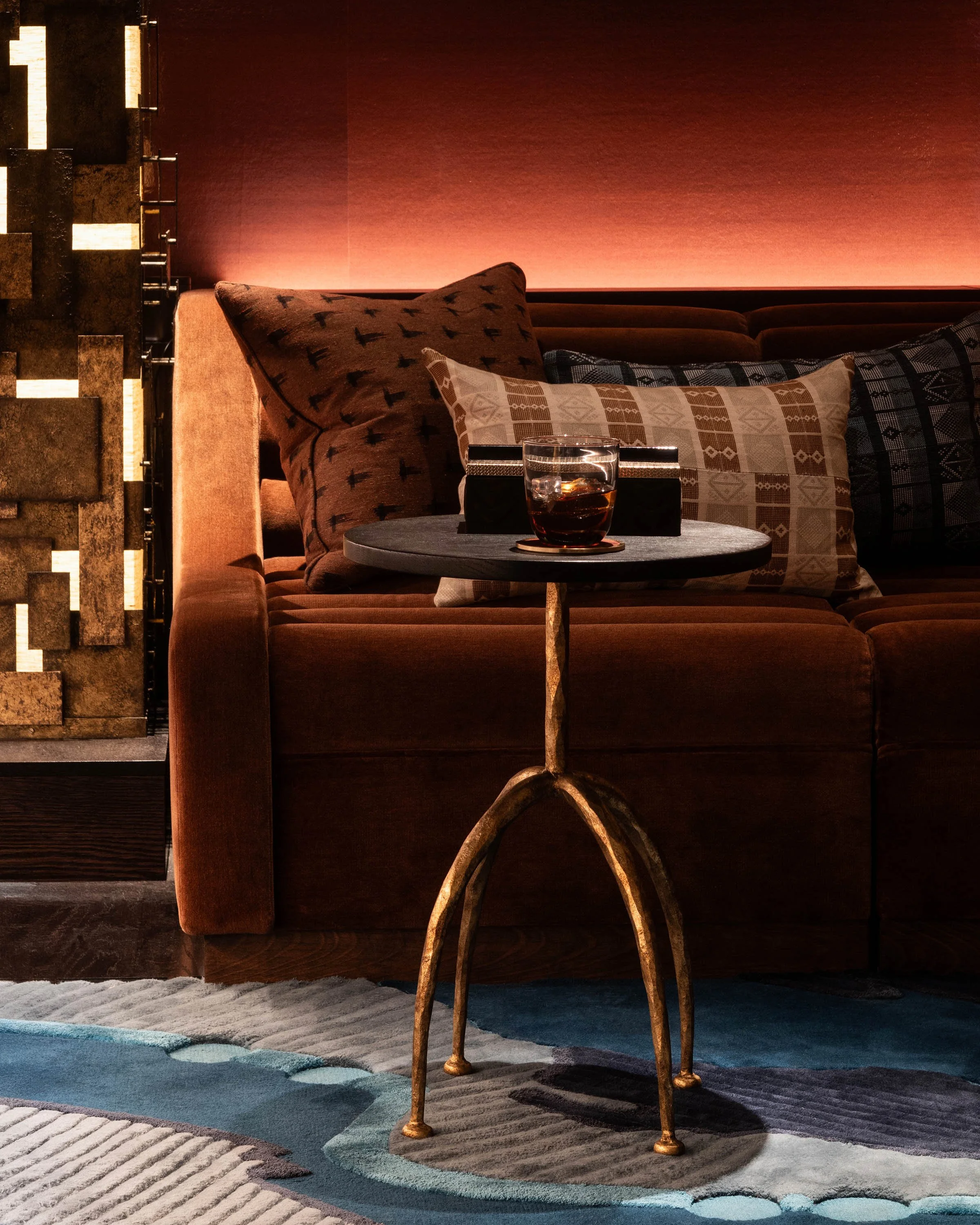 A cozy living room corner with a brown velvet sofa, decorative pillows, a small round side table with a glass of dark beverage, and a patterned rug.