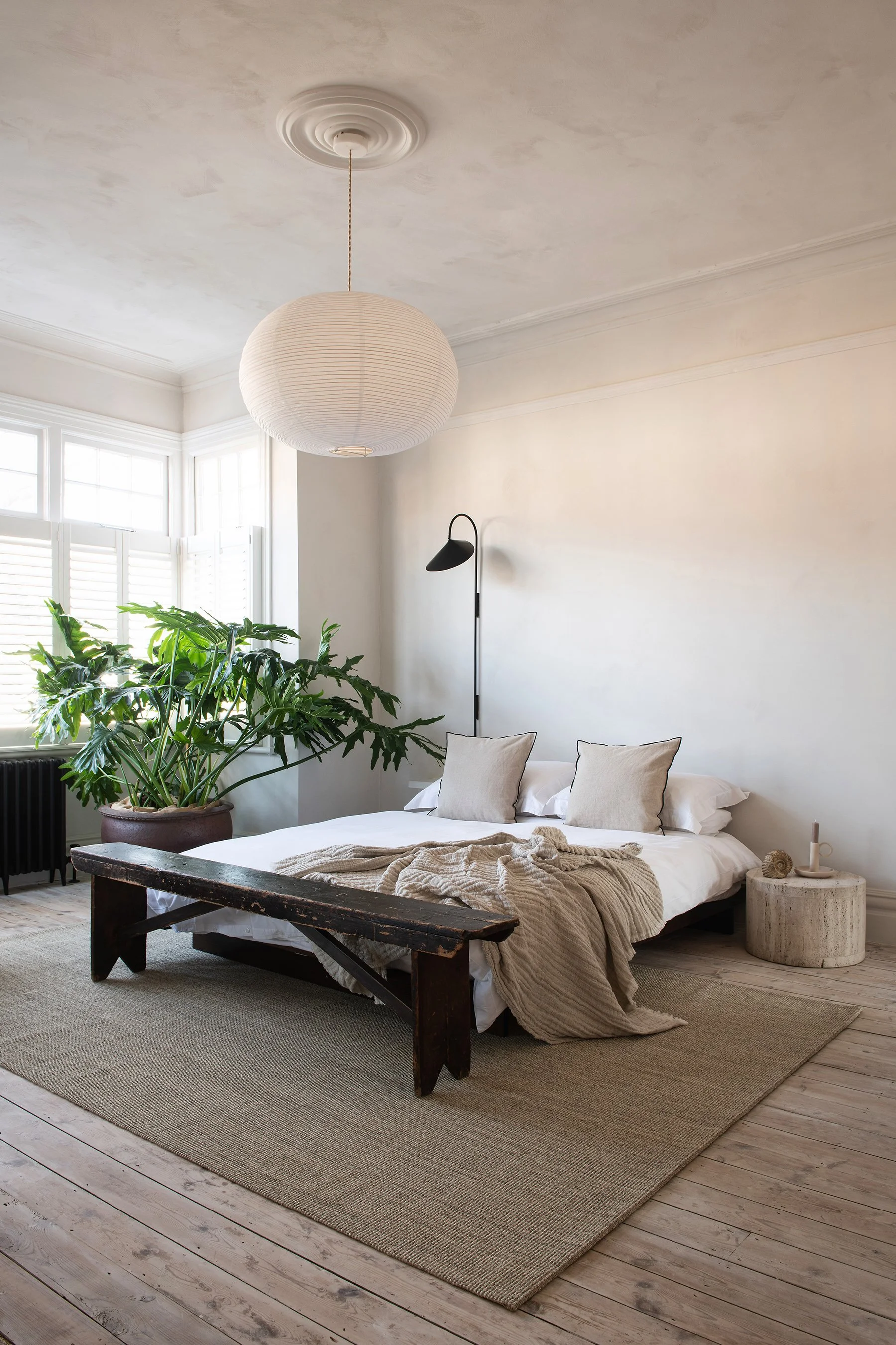 Brightly lit bedroom with a large green plant, a bed with white linens and beige pillows, a rustic wooden bench at the foot of the bed, a small round stone side table, and modern black wall-mounted reading light, with a paper lantern ceiling fixture.