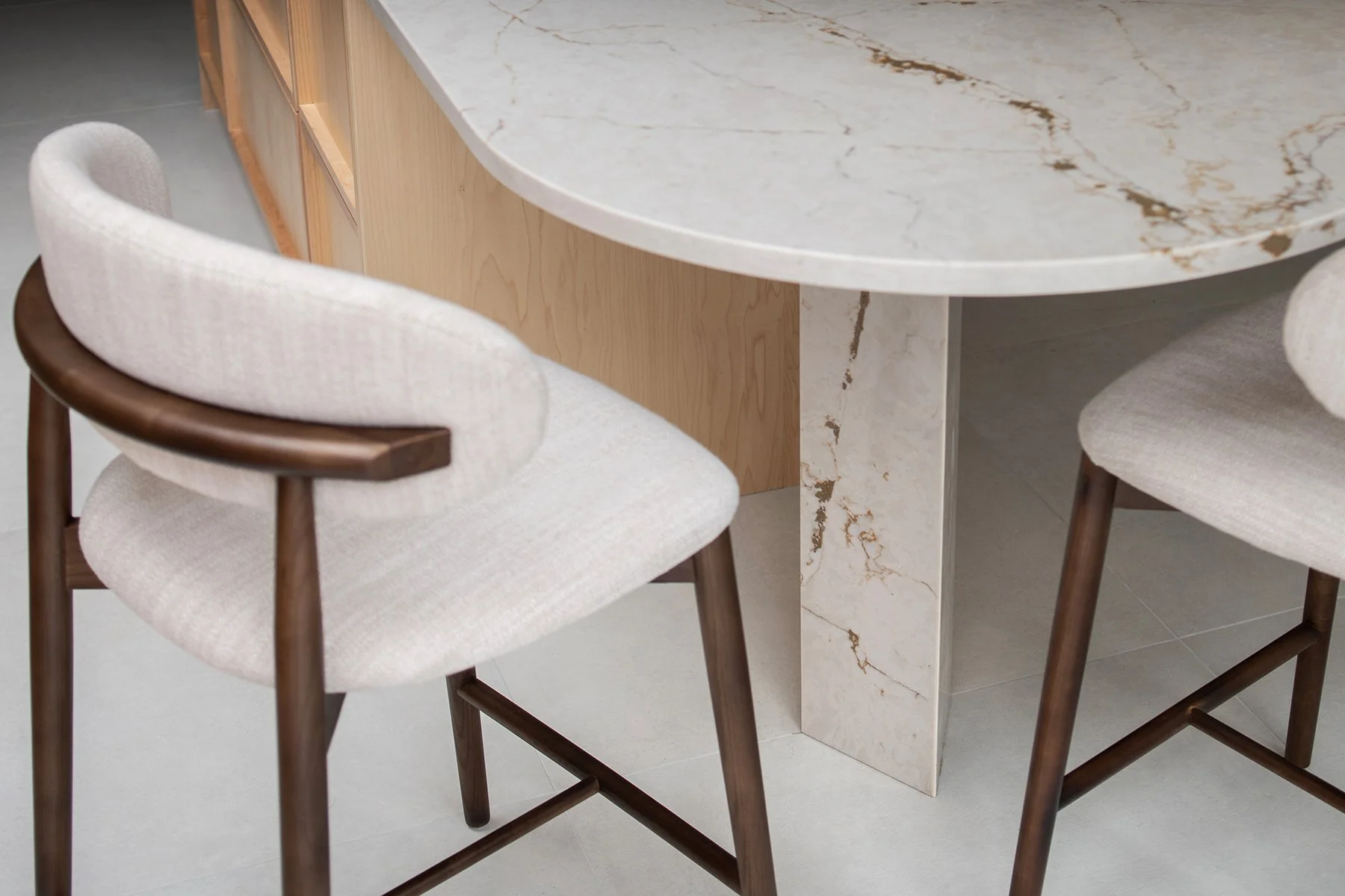 Close-up of a marble dining table with two light-colored upholstered chairs with wooden armrests and tapered legs.