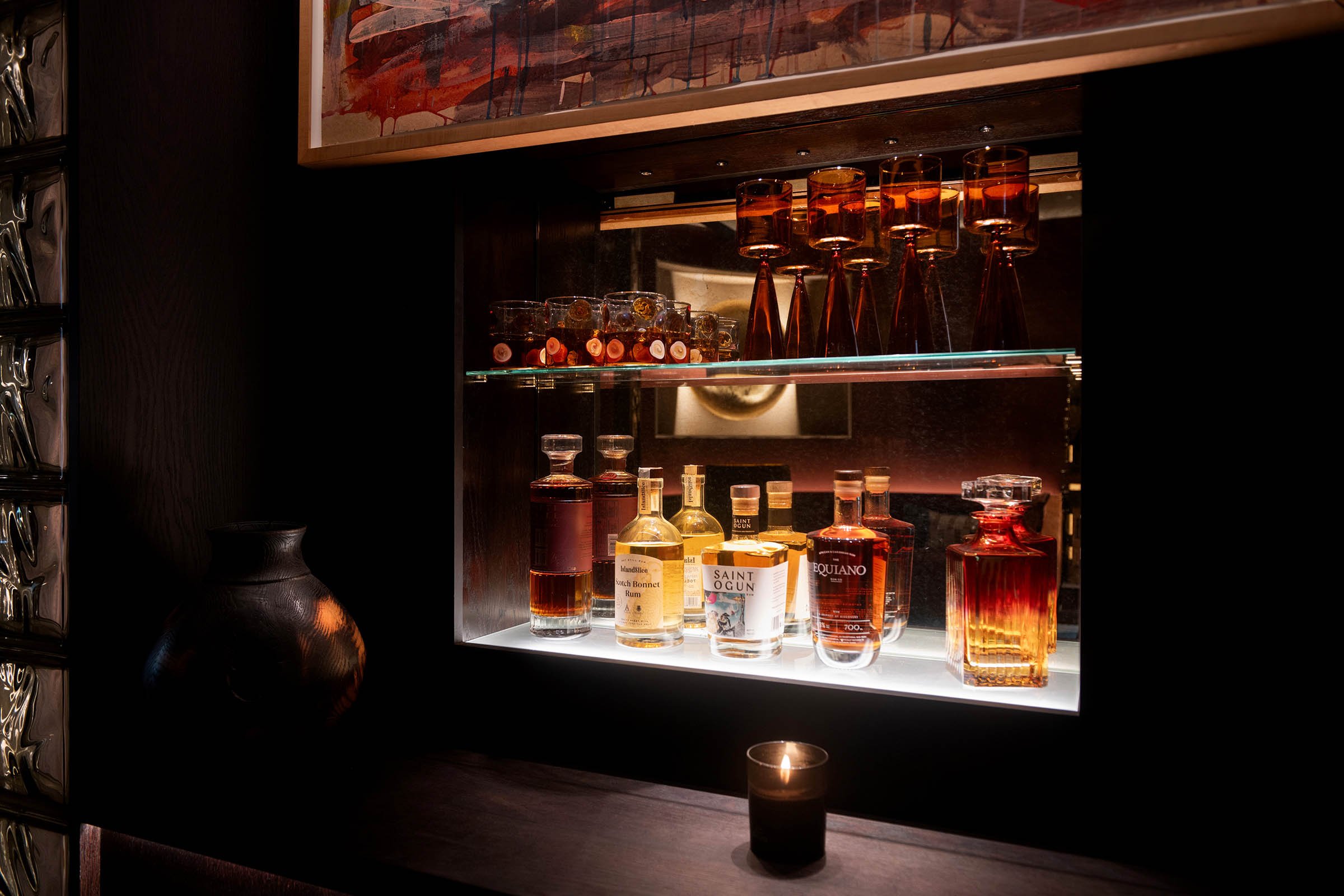 A backlit display of various bottles of liquor, including whiskey, rum, and brandy, along with cocktail glasses, on glass shelves in a dimly lit bar or lounge.