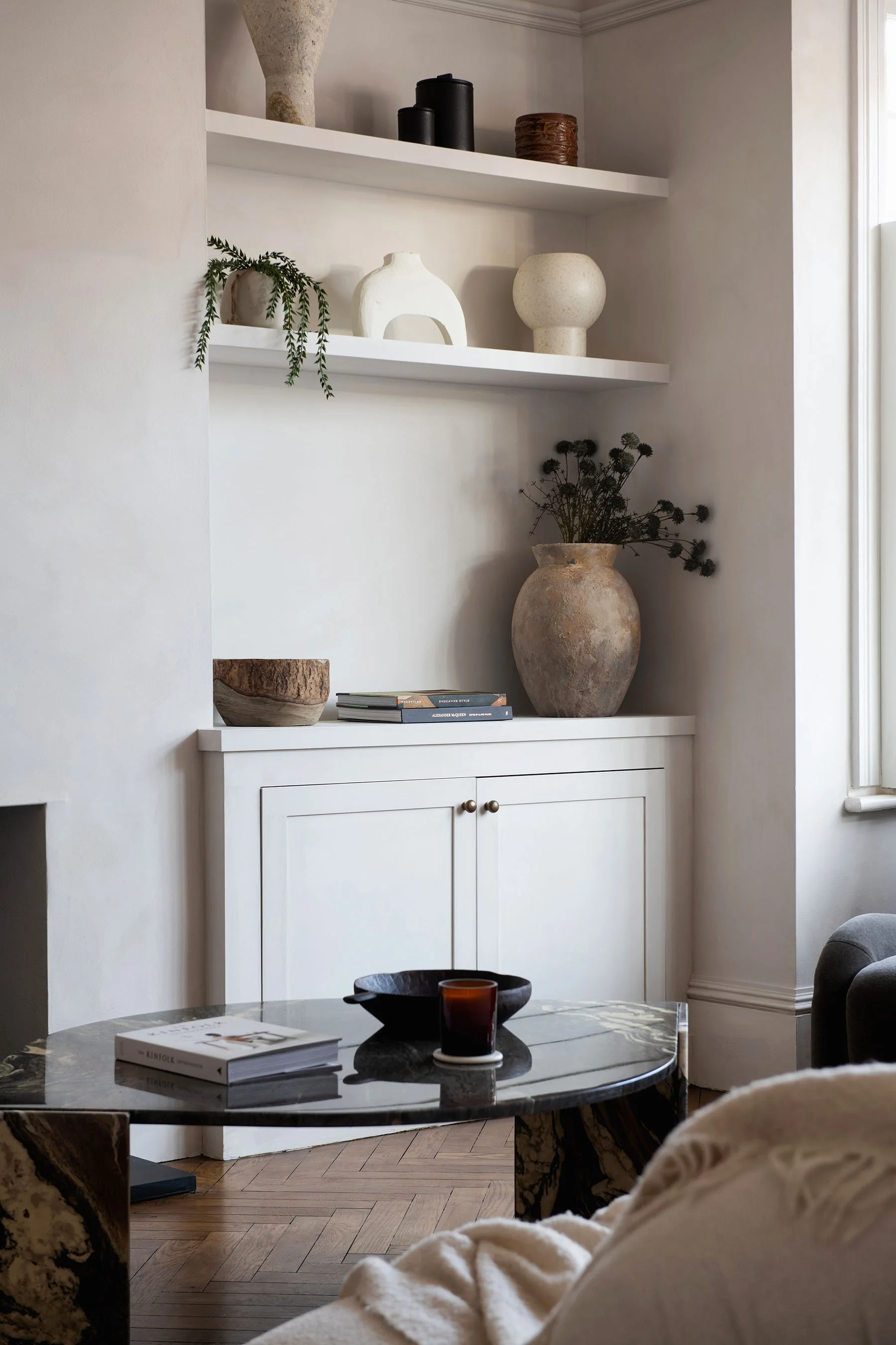 A living room corner with a black marble coffee table, a book, and a glass candle holder. White cabinetry with decorative pottery and books. Open wall shelves with vases, decor pieces, and plants. A window provides natural light.