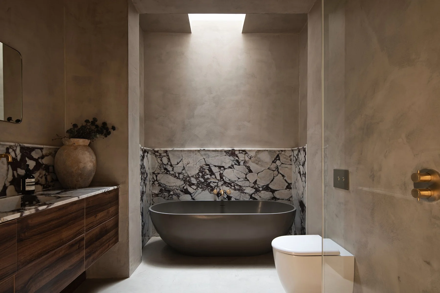 Modern bathroom with a freestanding black bathtub, marble accents, concrete walls, a toilet, a wooden vanity with a marble top, a vase with dried flowers, and a skylight overhead.
