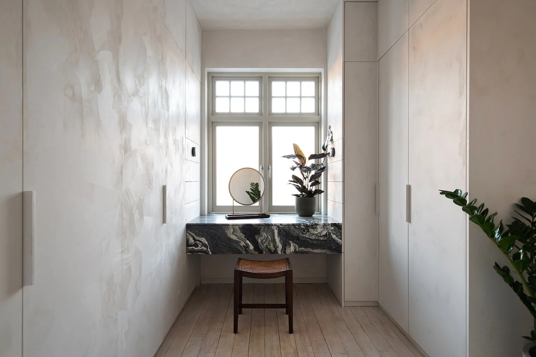 Small room with a window, a marble vanity table, a mirror, a potted plant, a stool, and white cabinets.