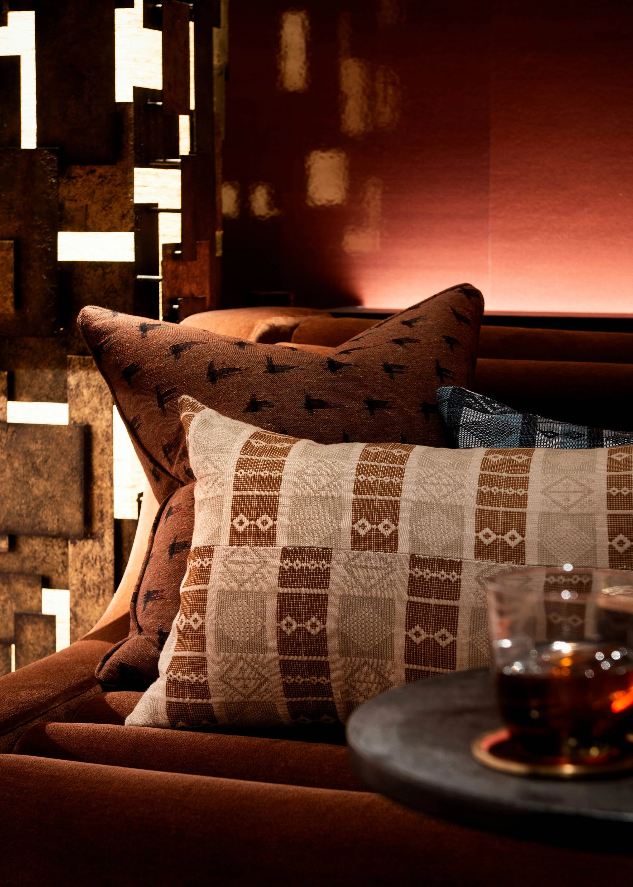 A cozy interior with a dark brown sofa decorated with patterned pillows in shades of brown, beige, and black, next to a small round table with a glass of red wine, and a textured wooden headboard or room divider in the background, illuminated by warm