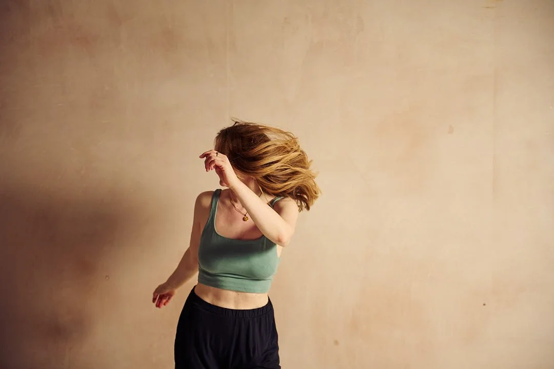 A woman with red hair wearing a green sports bra and black shorts, turning her head to the side with her hair flowing, against a plain beige wall.