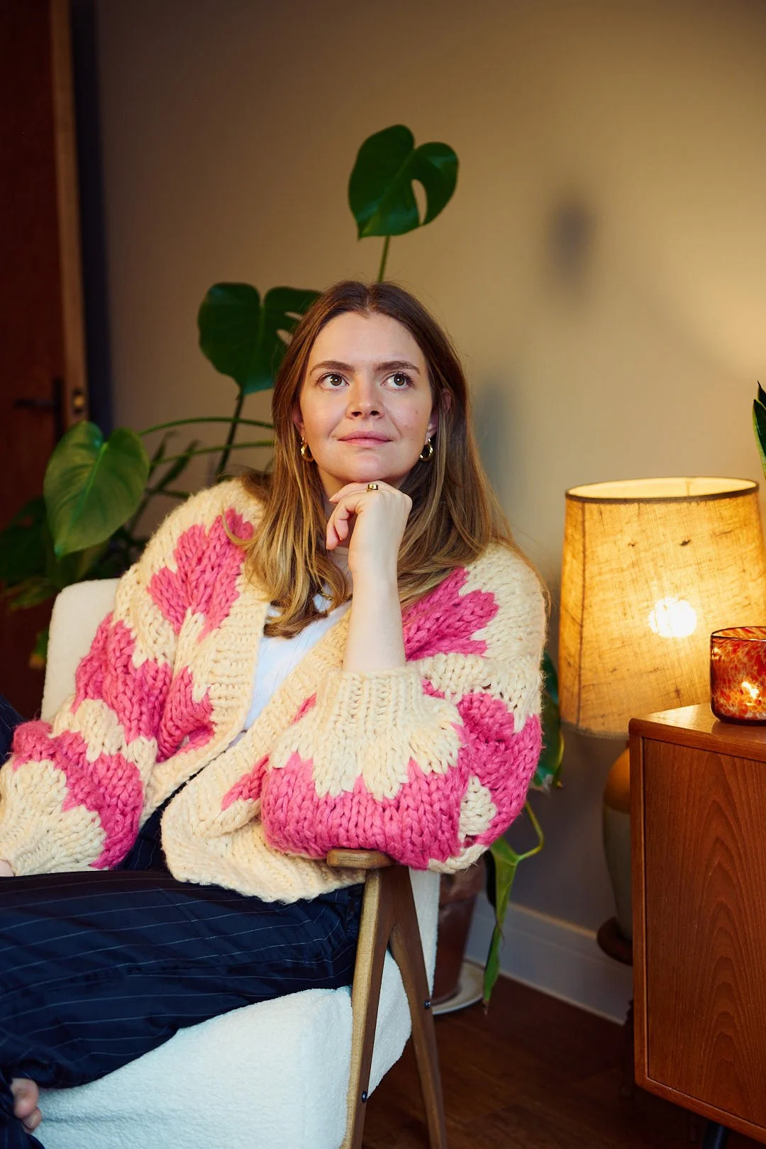 A woman with light brown hair, sitting on a white chair, wearing a chunky knit sweater with pink and cream patterns, resting her chin on her left hand, looking thoughtfully upward. Behind her are large green houseplants, a warm lit table lamp, and a small decorative candle on a wooden side table.