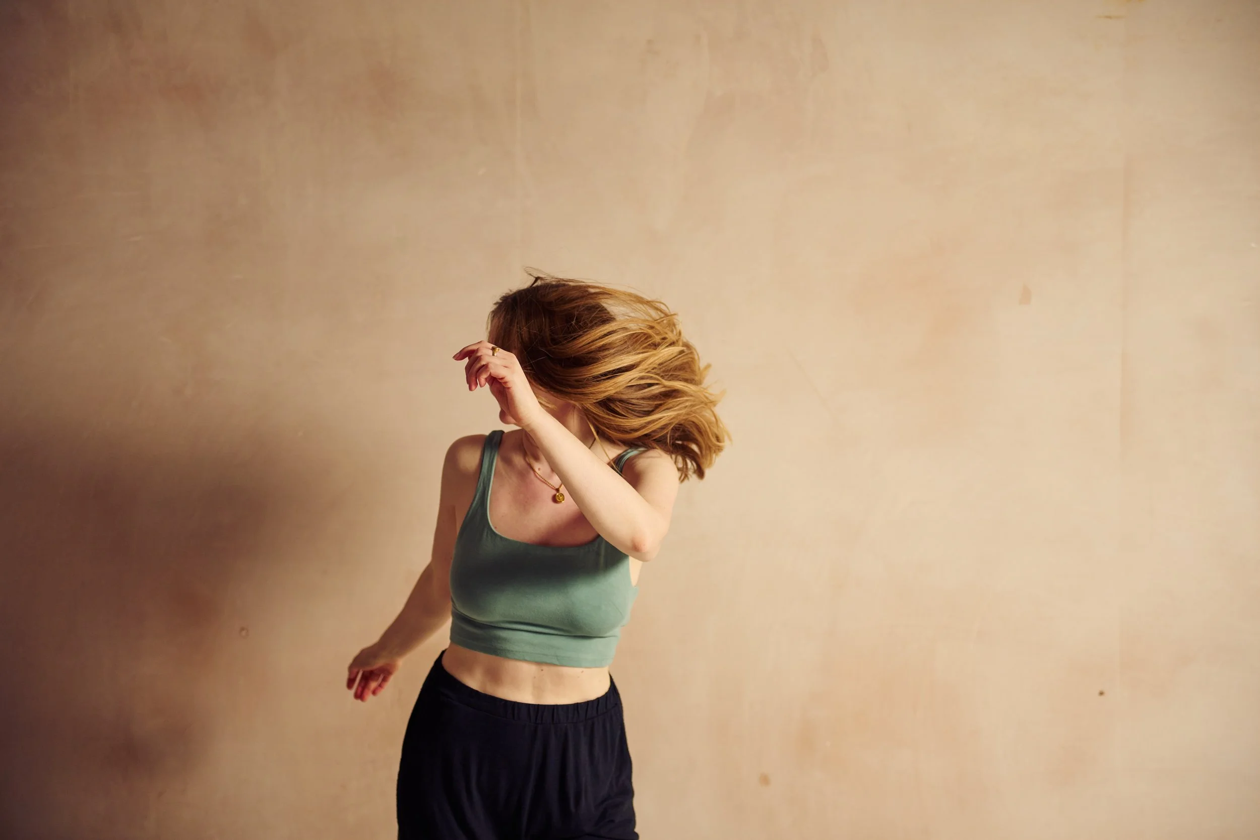 A person with red hair, wearing a green tank top and black pants, is standing against a plain beige wall. They appear to be in motion, with their head tilted down and one arm raised, covering part of their face.