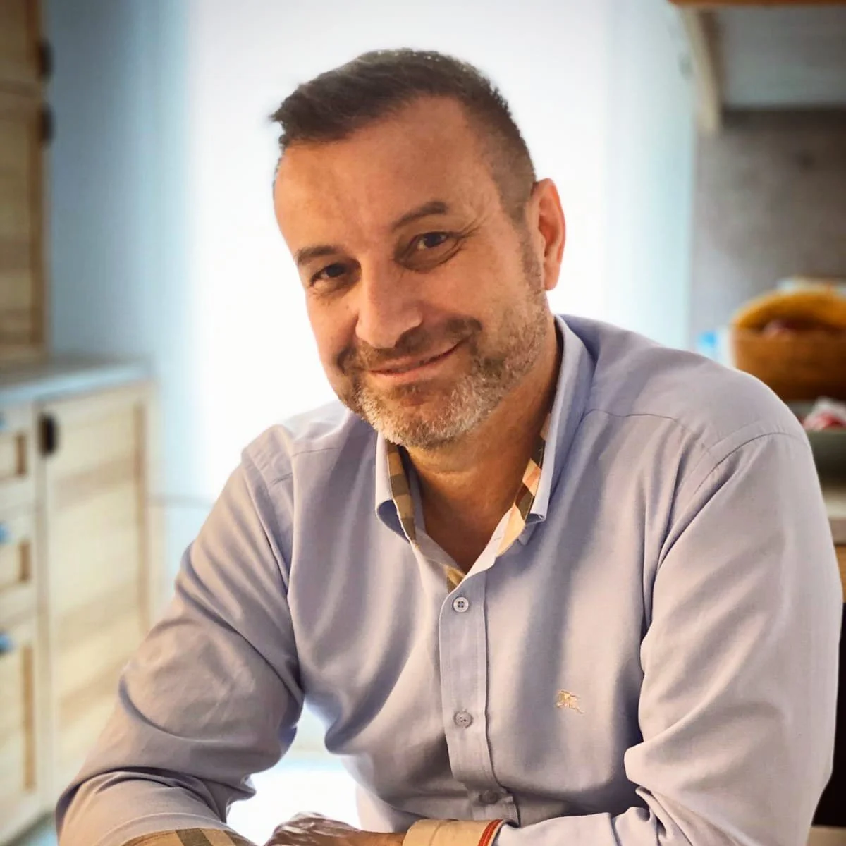 A smiling man with short brown hair and a beard, wearing a white button-up shirt, sitting indoors with a neutral background and a door visible in the background.