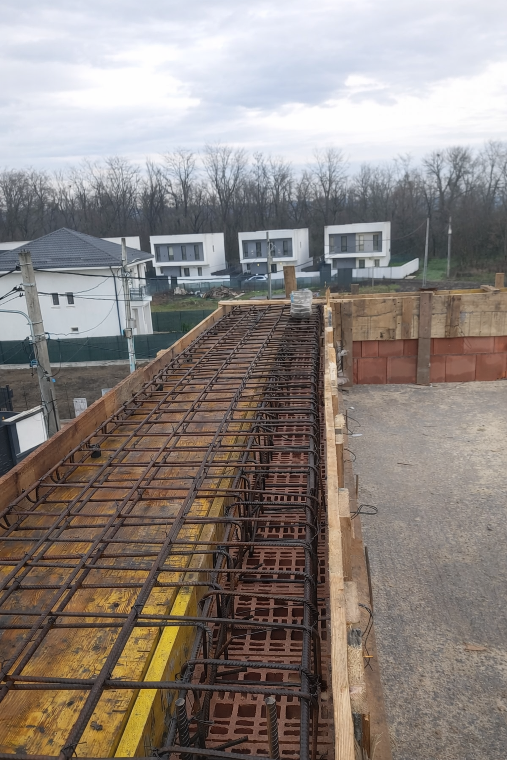 Building construction with steel rebar framework on the top floor, wooden formwork along the edge, and neighboring modern houses under a cloudy sky.