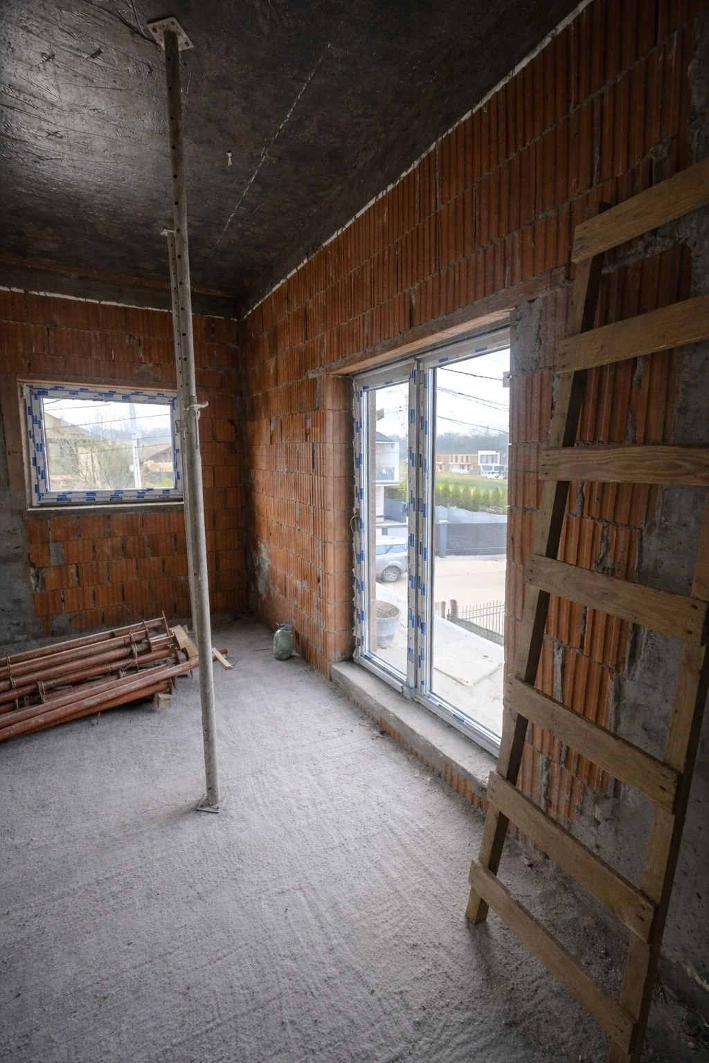 Interior of a house under construction with brick walls, a ladder, construction materials, and a window with a view outside.