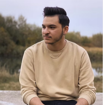 A young man with dark hair and a mustache sitting outdoors near a body of water, wearing a beige sweatshirt and a small chain necklace.