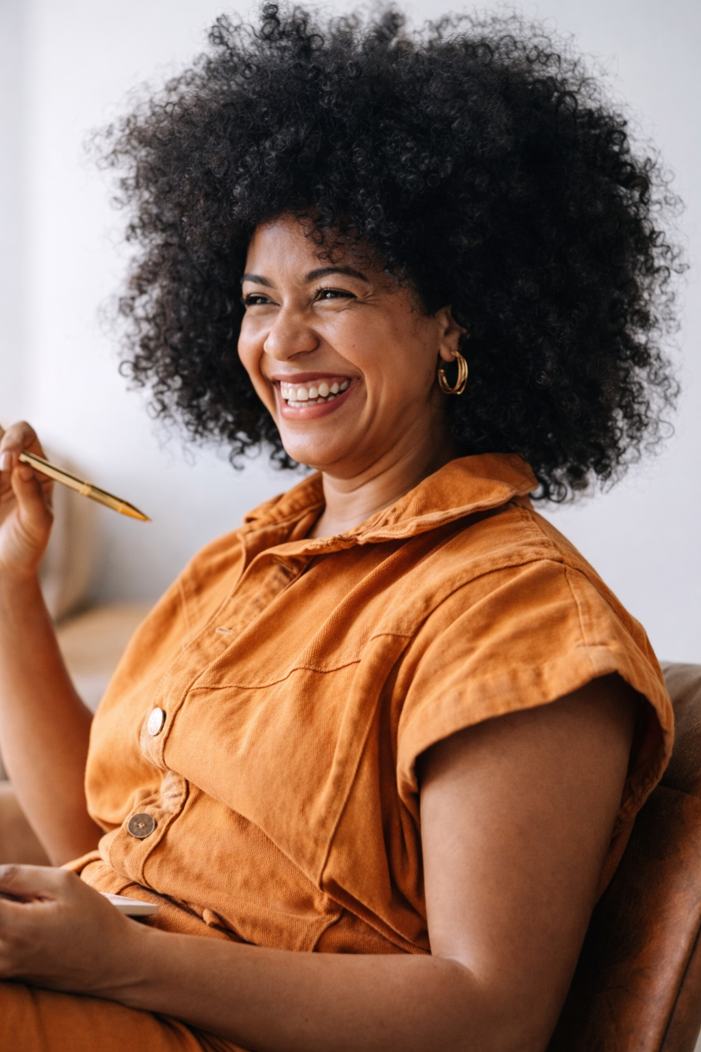 A woman with a big curly afro hairstyle, wearing gold hoop earrings and an orange short-sleeved button-up shirt, smiling and holding a pen in her right hand.
