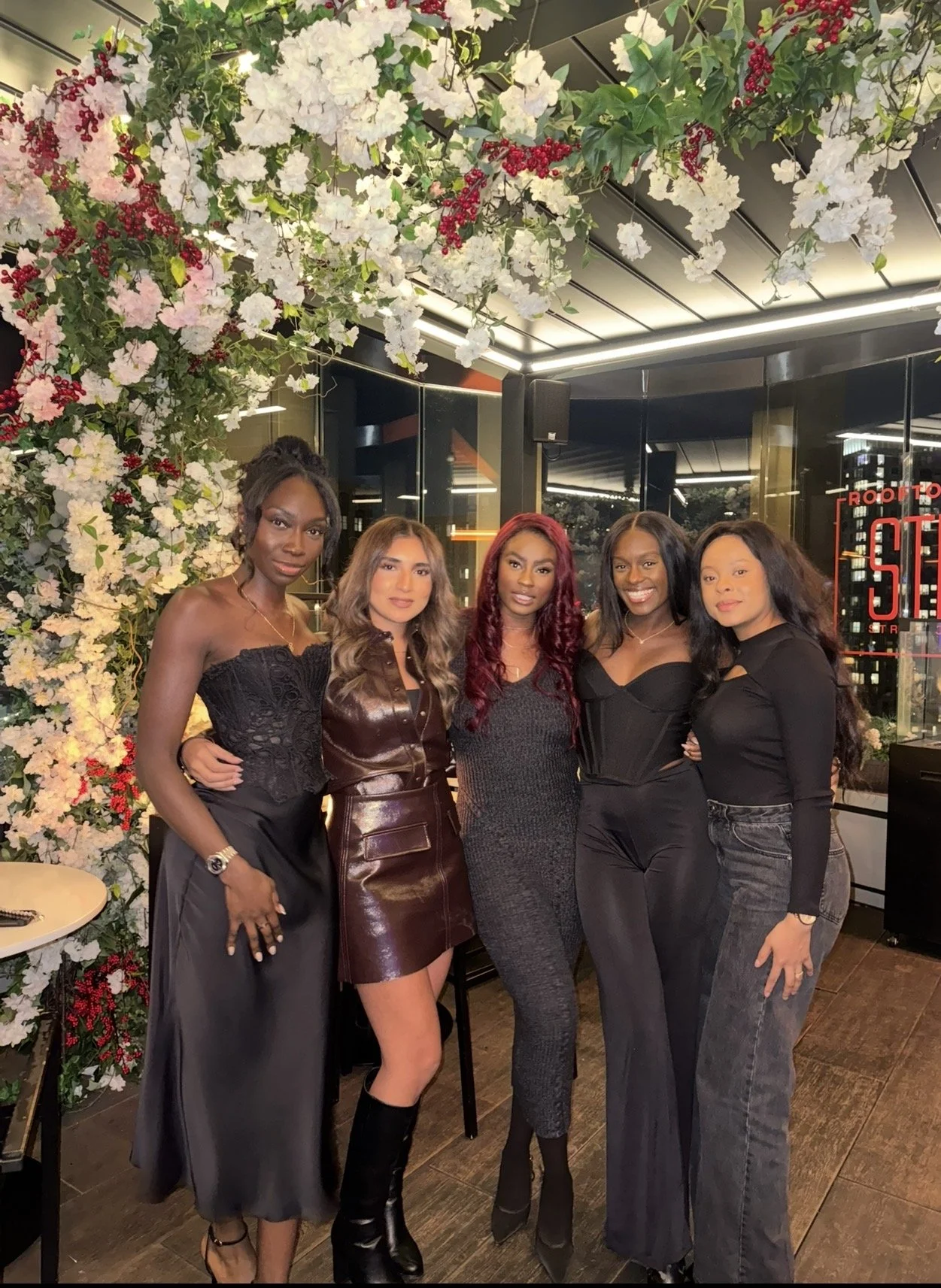A group of five women dressed in fashionable attire, standing together in an indoor setting with floral decorations overhead and modern decor visible in the background.