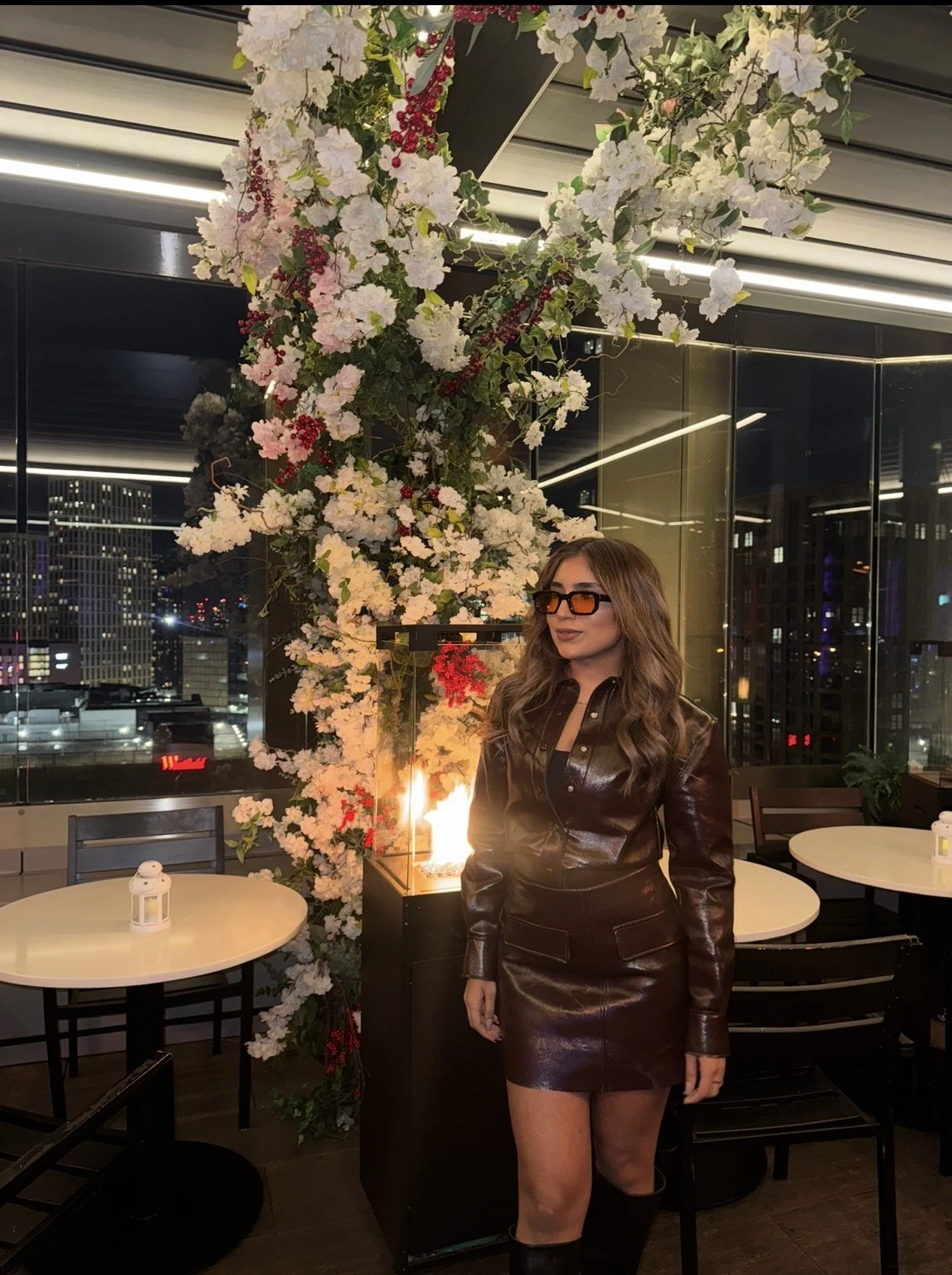 A woman in a leather outfit and sunglasses standing next to a lit fireplace and floral arrangement in an urban restaurant with city lights visible outside.