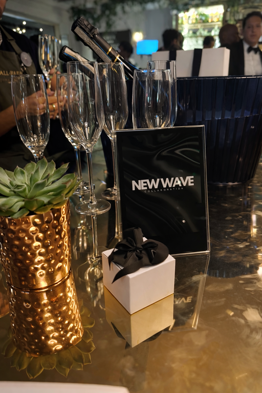 Celebration setup with empty champagne flutes, a succulent plant in a textured bronze planter, a small gift box with a black ribbon, and a sign that reads 'NEWWAVE COLLABORATION' on a reflective table at an indoor event.