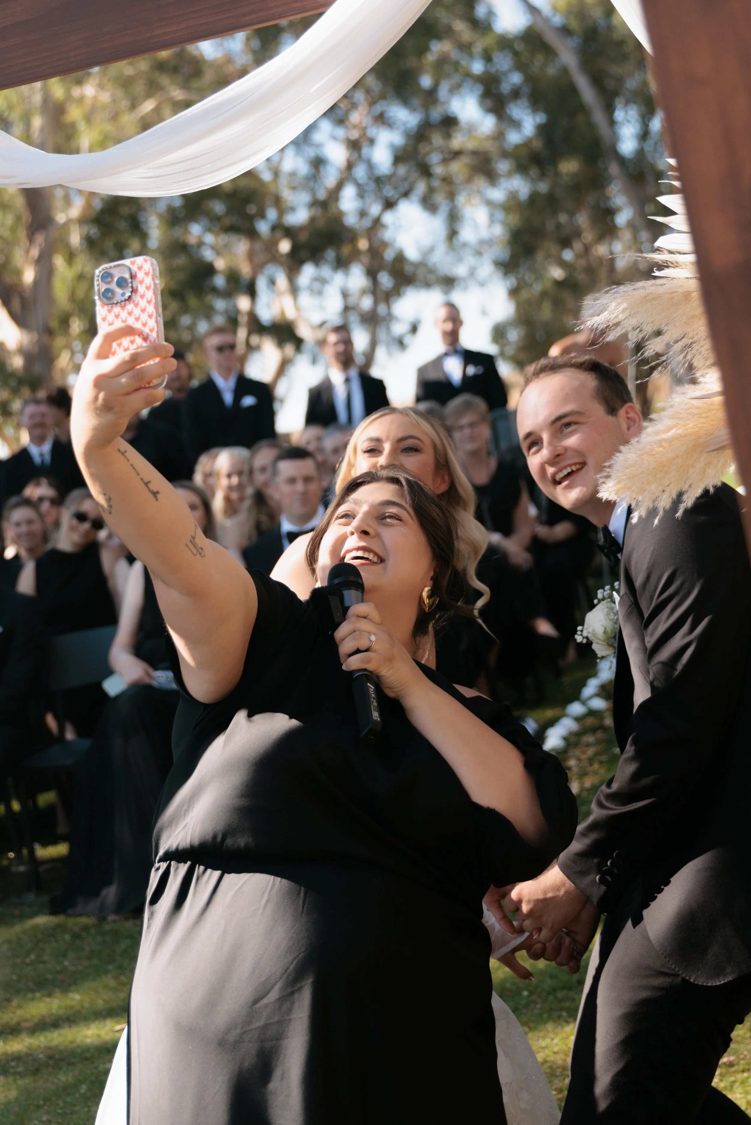 A bride and groom celebrate their wedding outdoors with friends and family. The bride is holding a microphone and taking a selfie with her guests, smiling happily. The groom is holding the bride's hand, also smiling. Everyone is dressed in formal attire, with trees and a white canopy visible in the background.