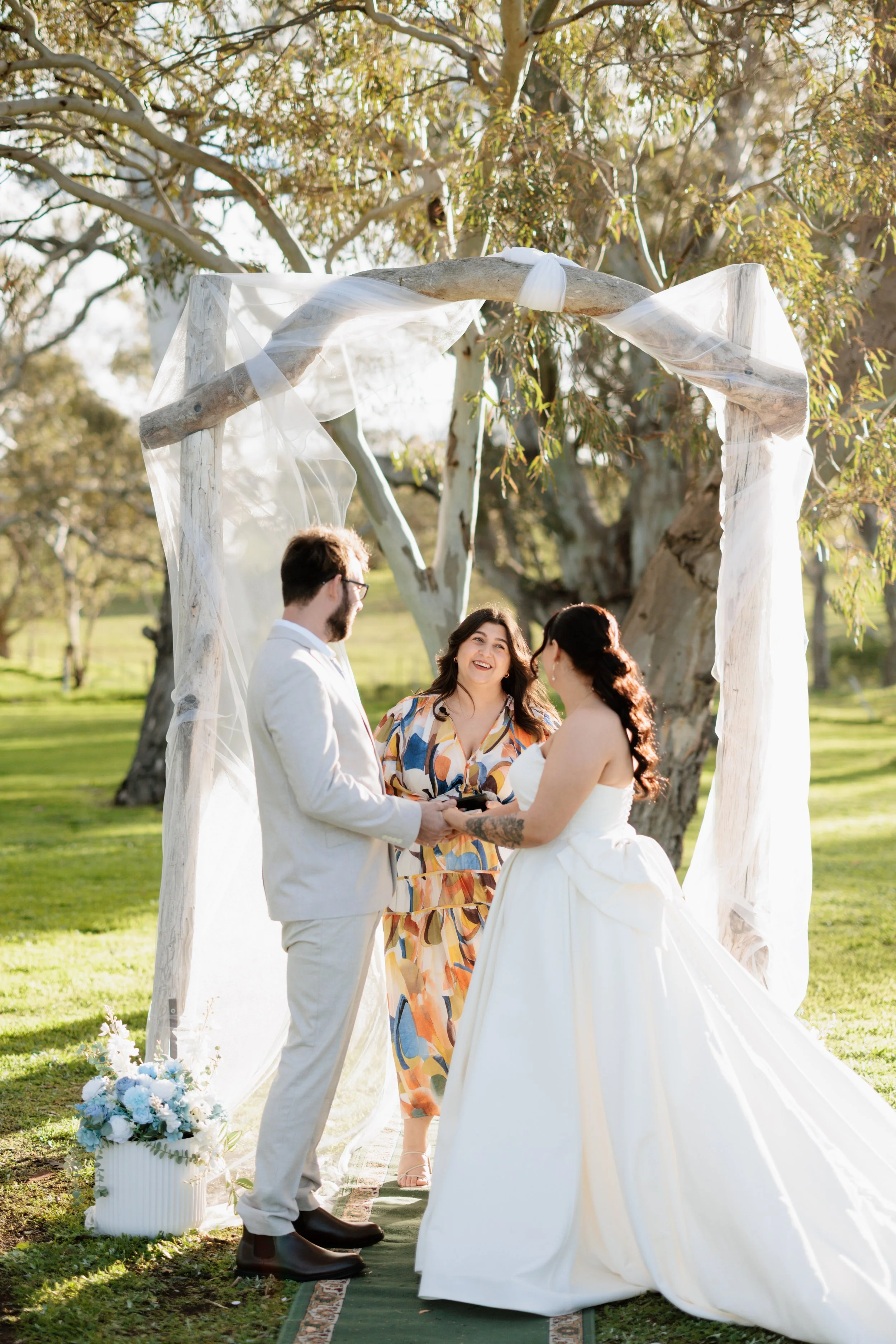 A wedding ceremony outdoors with a bride, groom, and officiant under a rustic wooden arch decorated with white fabric, in a green park with large trees.