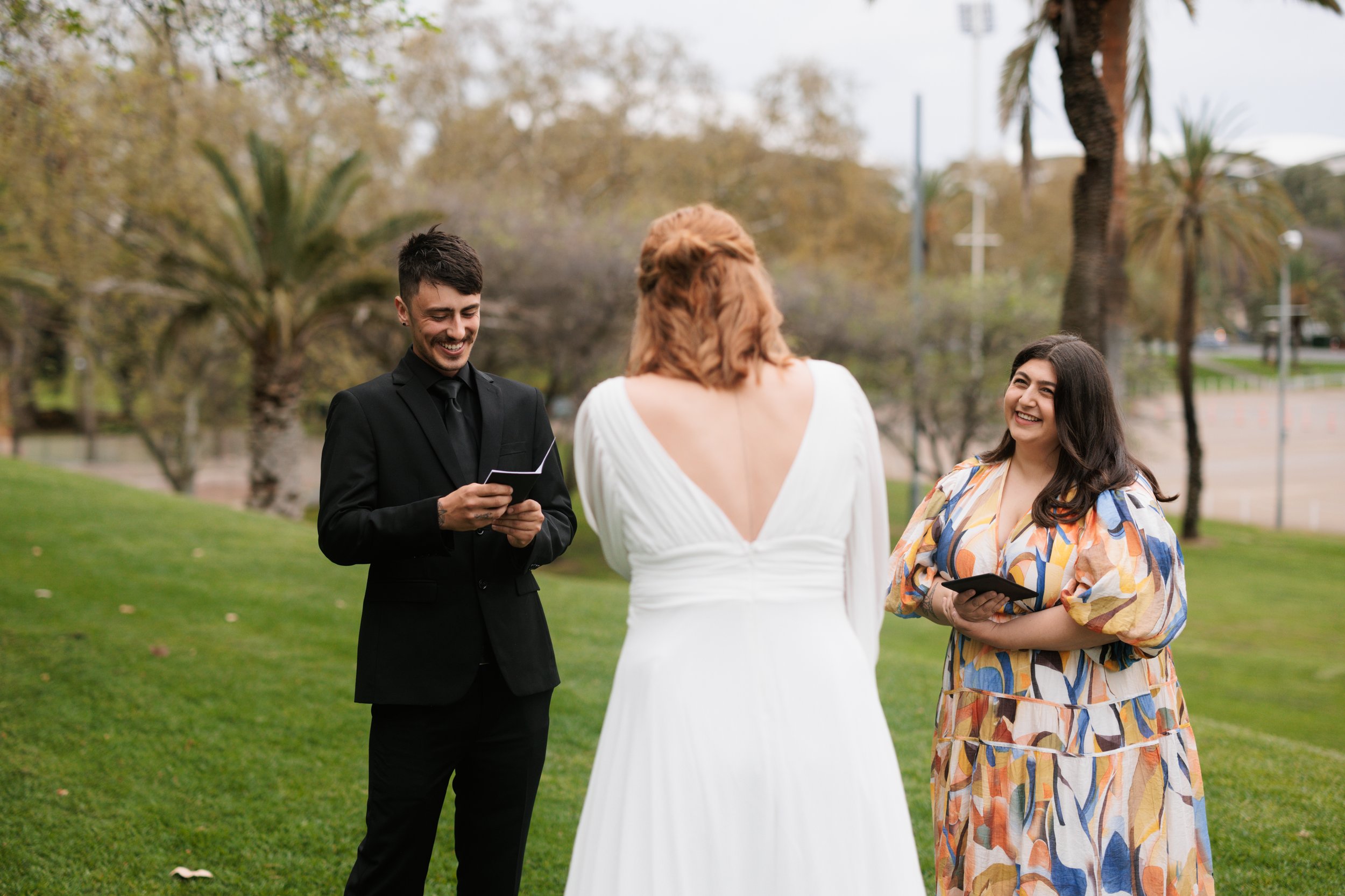 A couple getting married outdoors, with the officiant reading vows. The groom is wearing a black suit, and the bride has shoulder-length red hair and is dressed in a white wedding gown. An officiant stands between them, holding a booklet.