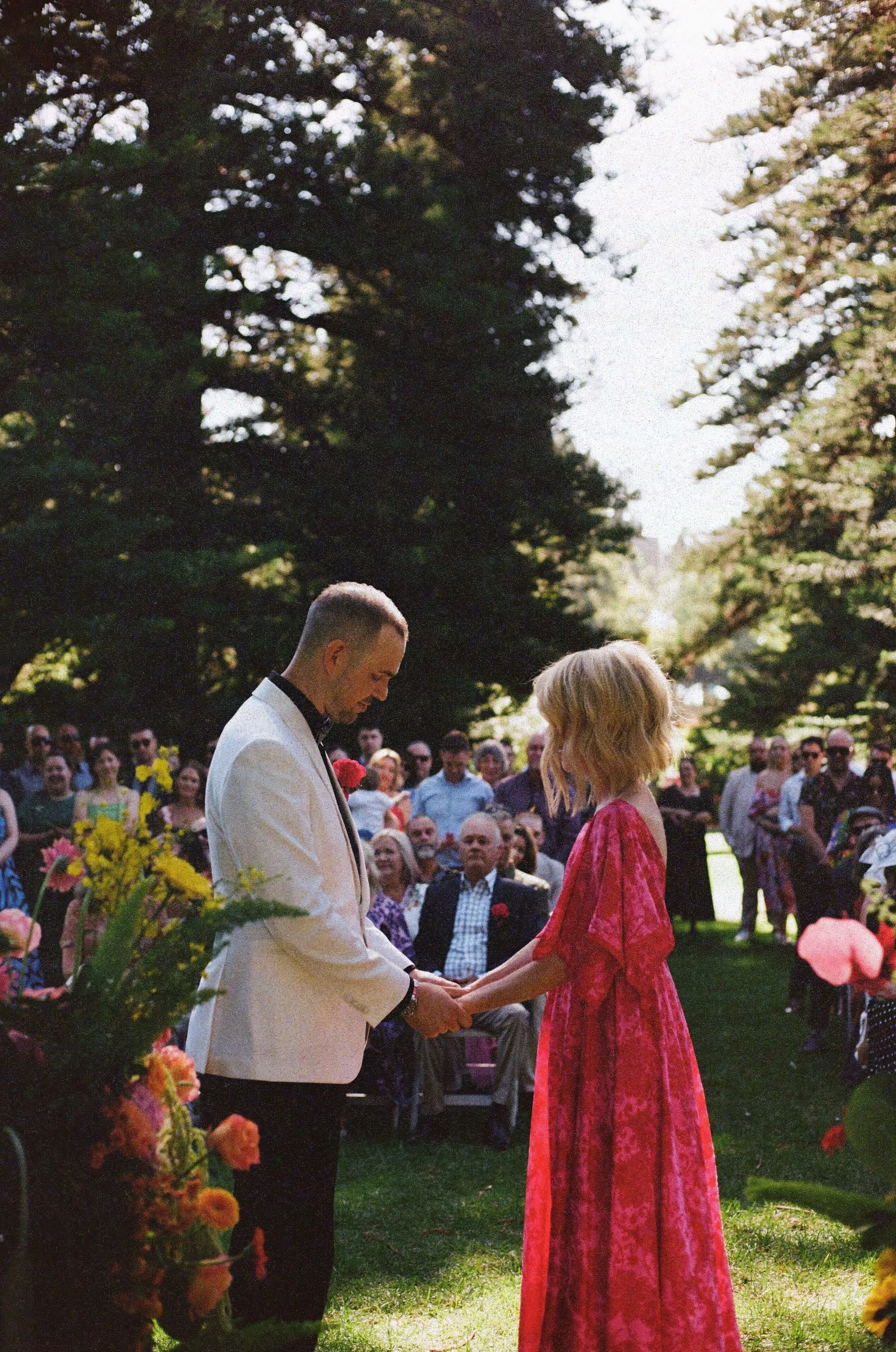 A wedding ceremony outdoors with a man in a white tuxedo and a woman in a long red dress holding hands, surrounded by seated and standing guests under trees.