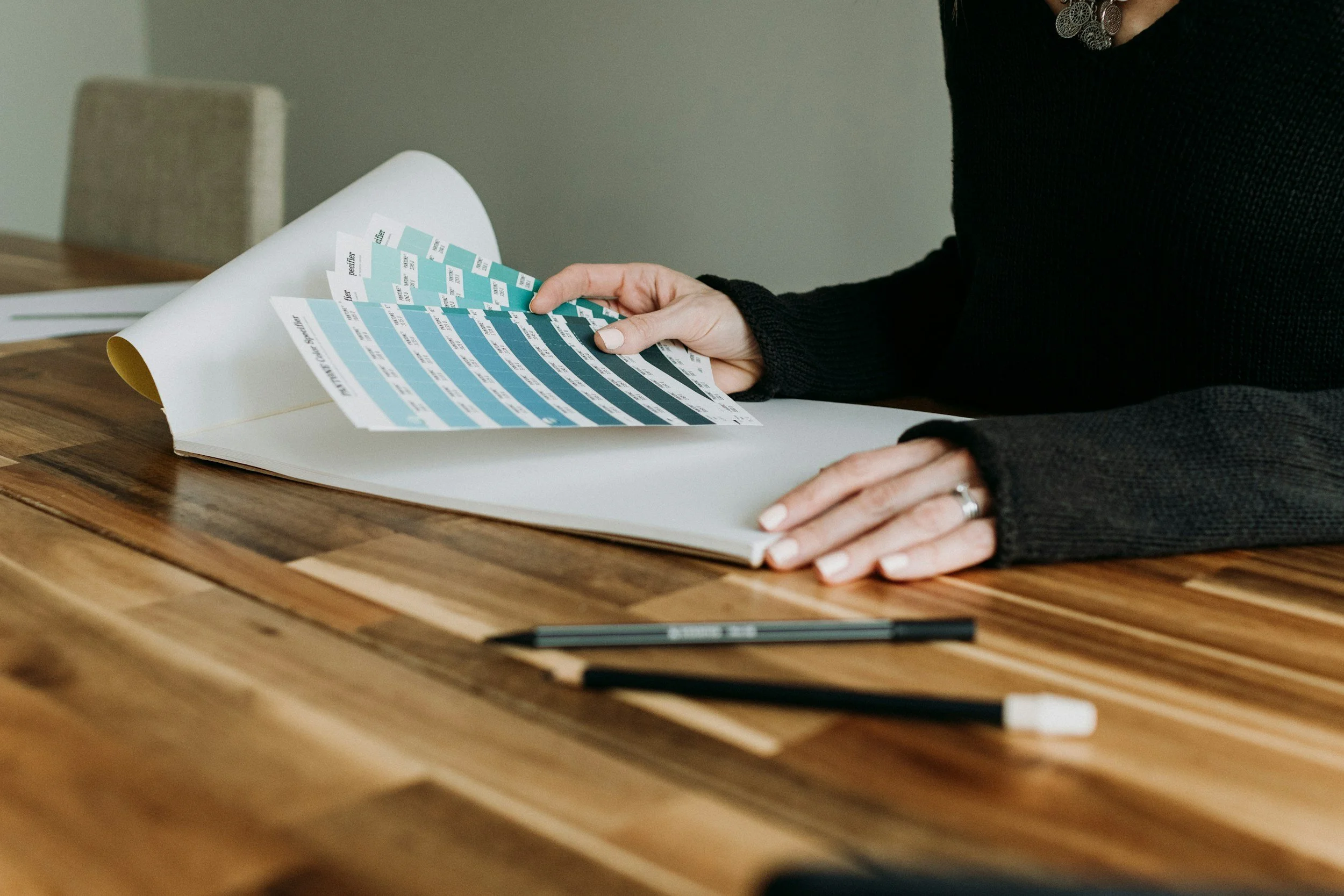 Person in black sweater flipping through color swatches or paint samples on a large blank page, with pens and papers on a wooden table.